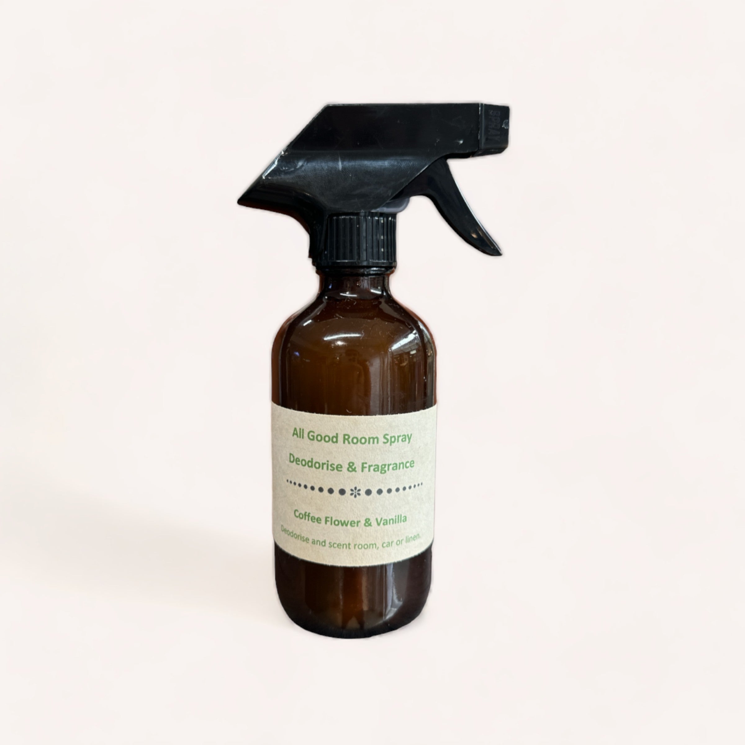 A brown glass spray bottle with a black trigger nozzle labeled "Coffee Flower & Vanilla Room Spray by All Good Bags, deodorizing freshener, for home and recent room, or car." against a plain.