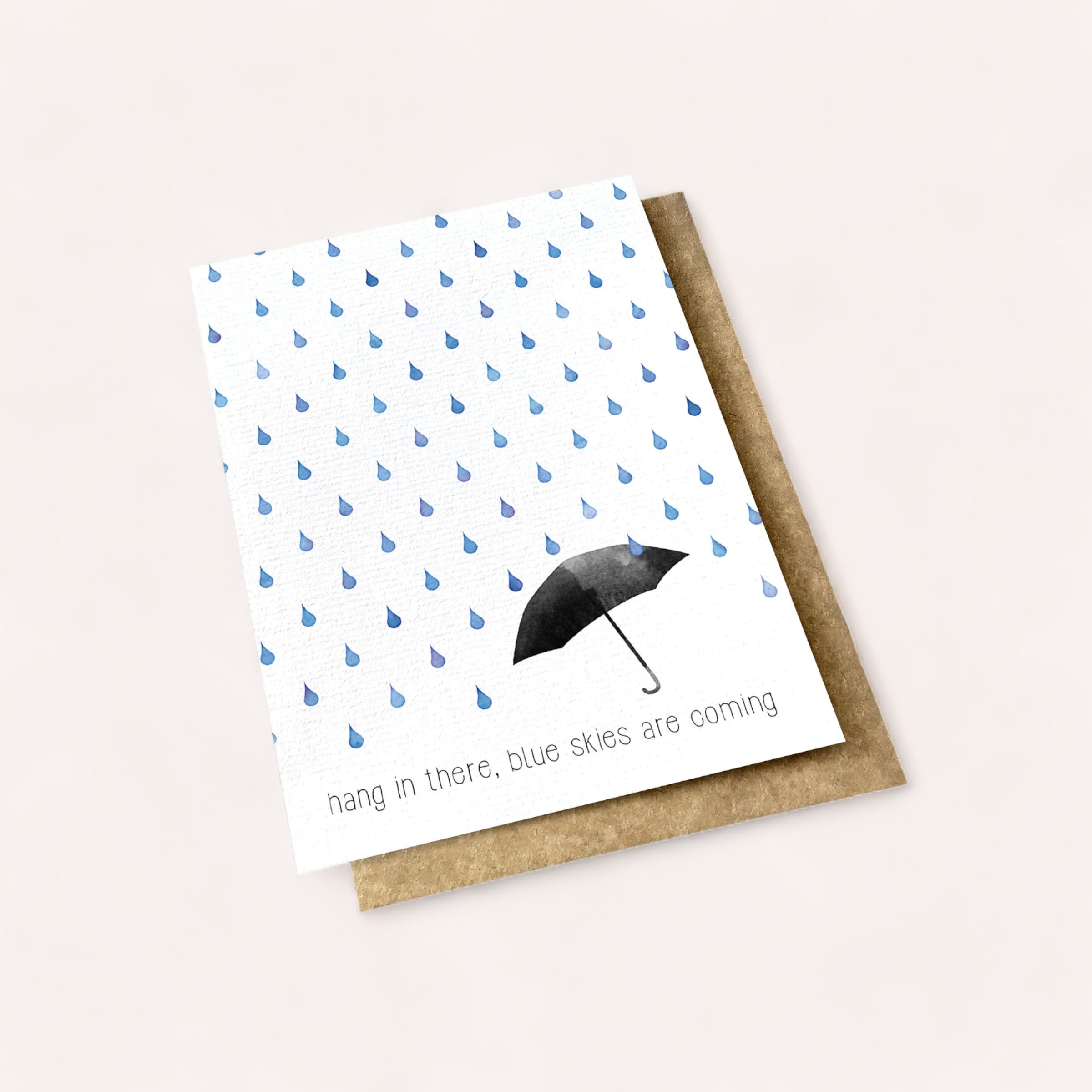 A "Blue Skies Are Coming Card" with a hopeful message, featuring a pattern of blue raindrops and a single black umbrella, with the text "hang in there, blue skies are coming." This product of New Zealand is by Ink Bomb.