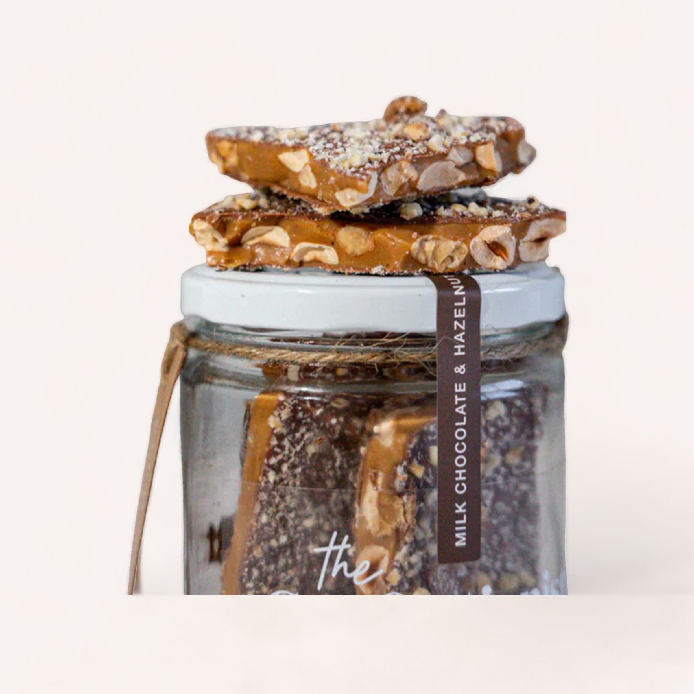 A glass jar labeled "Hazelnut Milk Chocolate Toffee" by The Confectionist filled with chocolate pieces, topped with two chocolate hazelnut bars sprinkled with sea salt, against a white background.