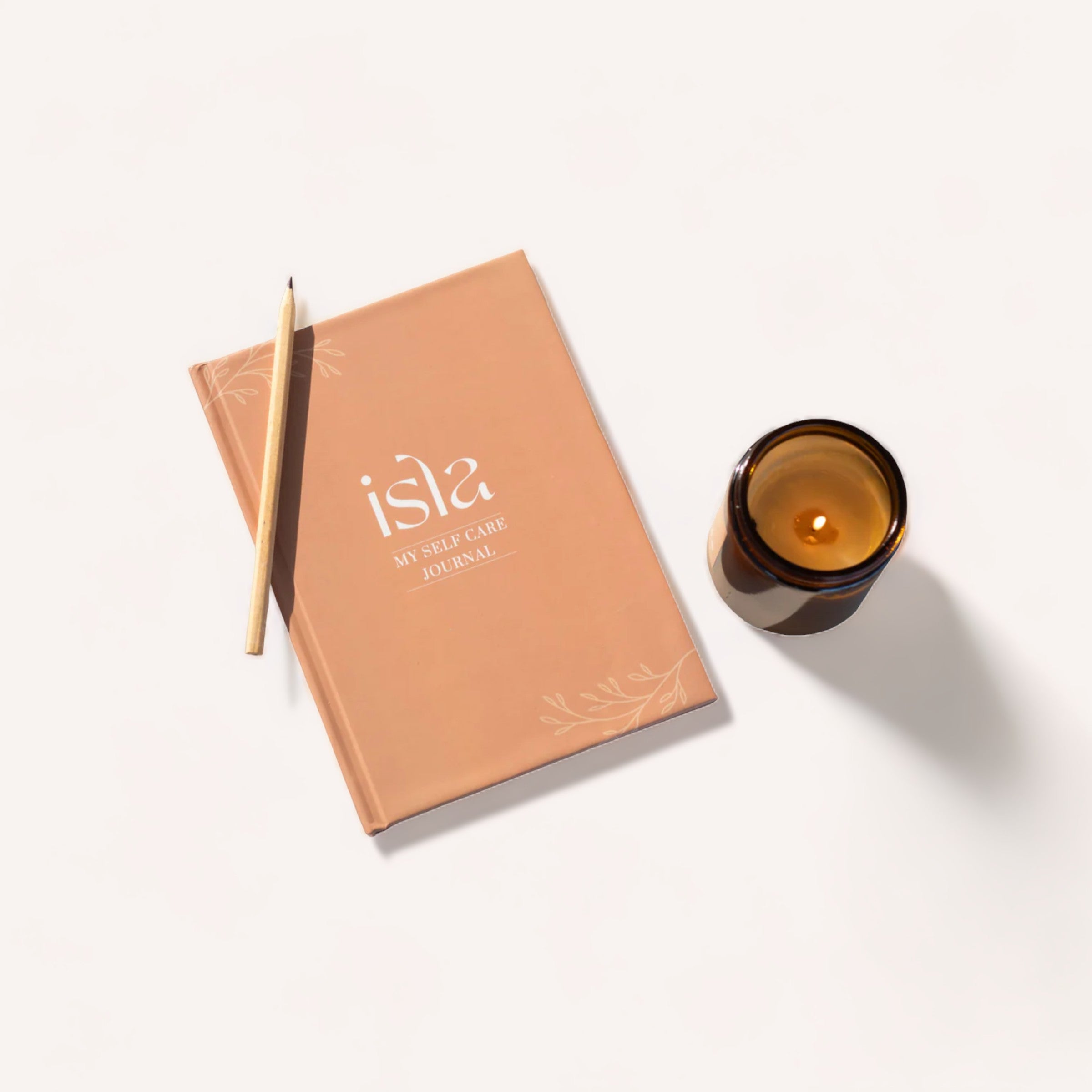 A peach-colored Skye self-care journal planner with a pencil on top lies next to a lit scented candle with a warm amber glow, all arranged on a clean white surface, suggesting a tranquil setting for mindfulness.