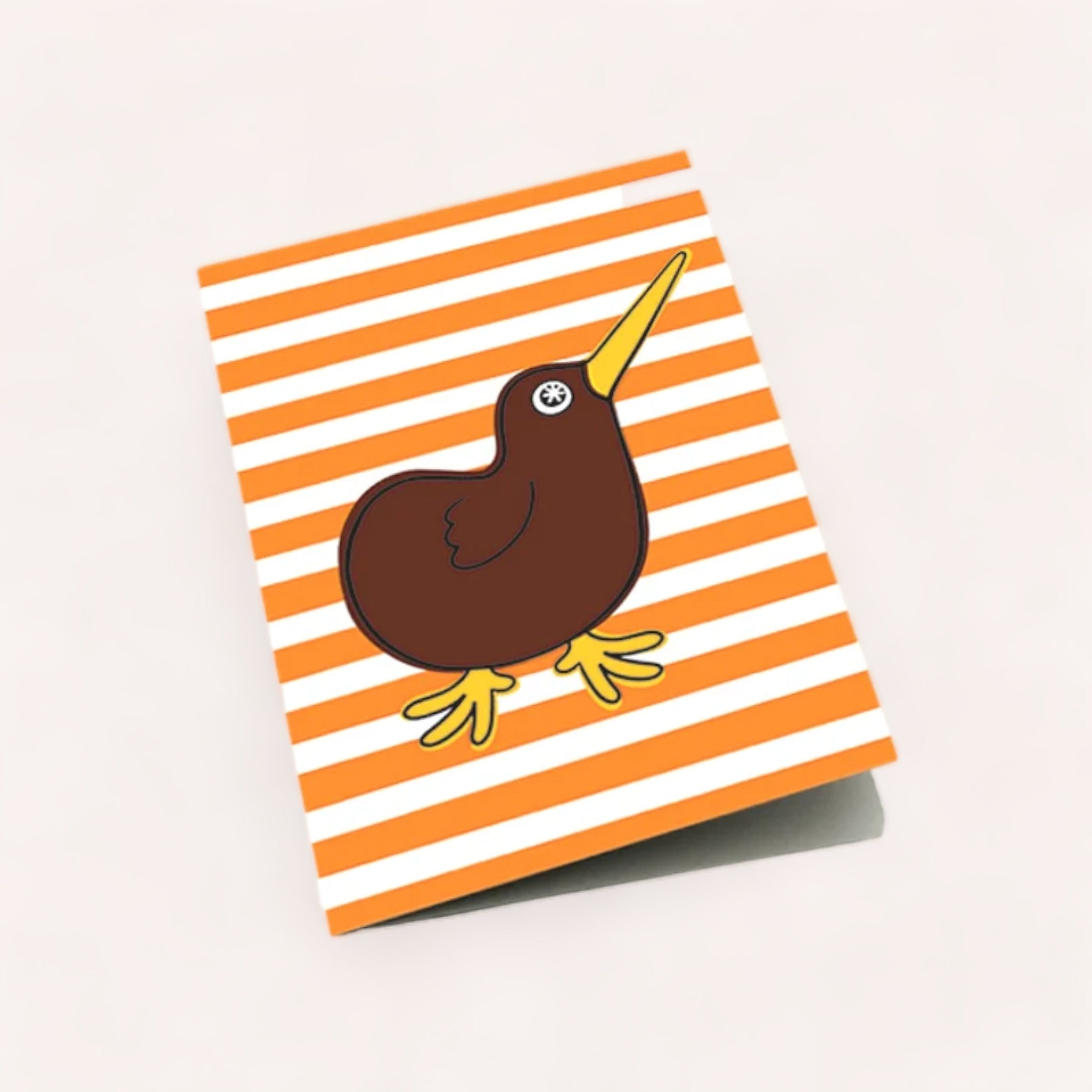 A whimsical sticker of a brown, cartoon-style bird with a long yellow beak and feet placed on a Kiwi Buddy Card by Design by Leonard lying on a light surface.