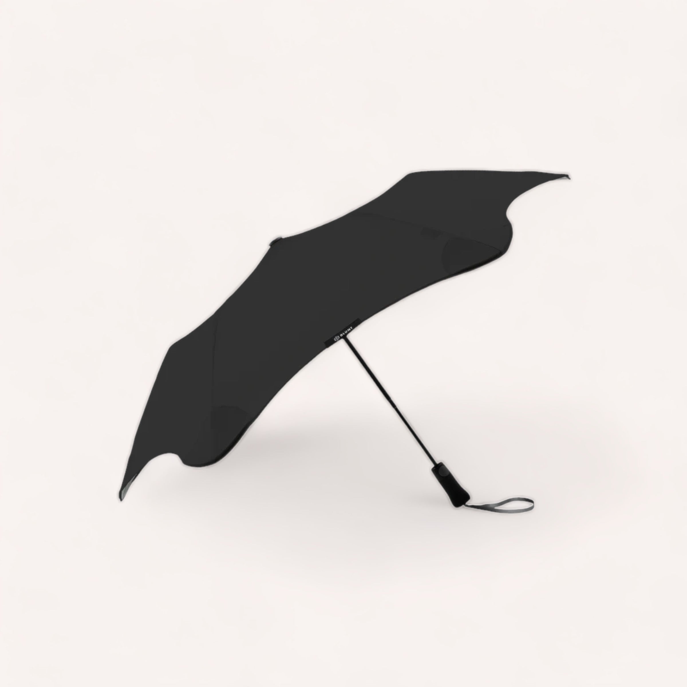 A sleek BLUNT Metro umbrella with an auto-open canopy opened on a white background, seemingly caught mid-motion as if tossed by a gentle breeze.