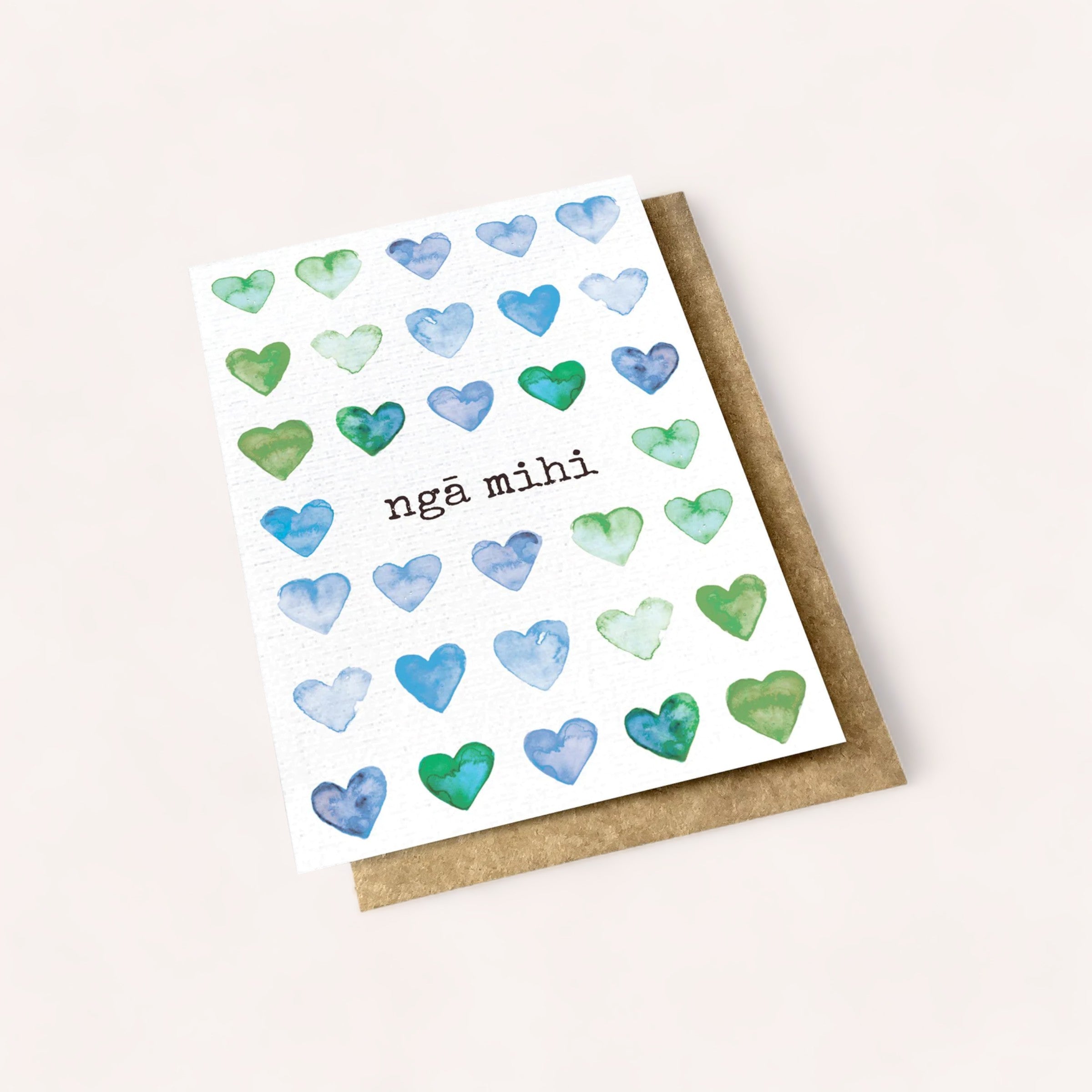 A Nga Mihi Card with watercolor hearts in shades of blue and green, featuring the words "nga mihi" on an off-white background, paired with a kraft paper envelope. This Product of Ink Bomb.