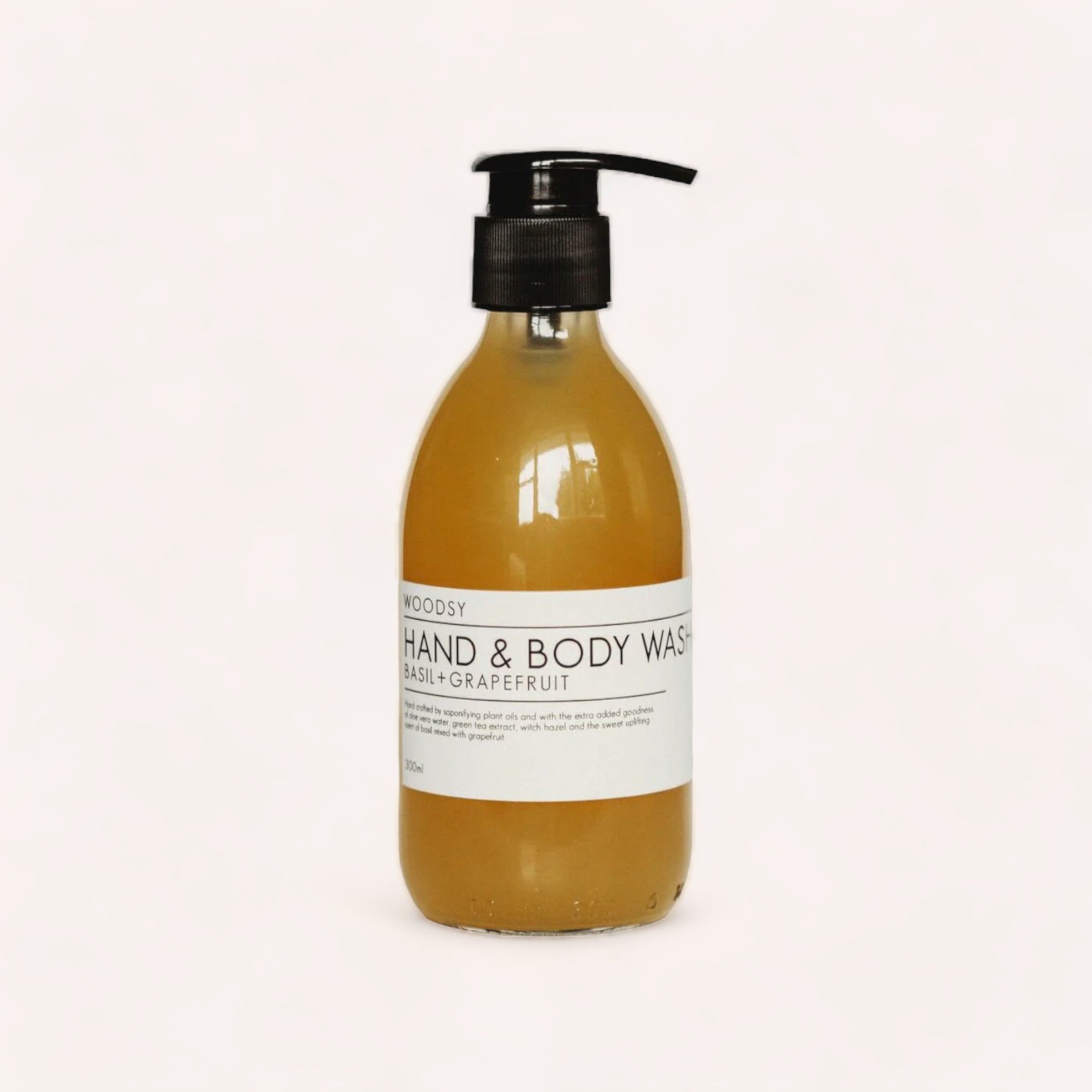 A pump bottle of Hand & Body Wash - Basil & Grapefruit by Woodsy Botanics with a label that indicates it's a woodsy, basil grapefruit-scented product containing organic oils.