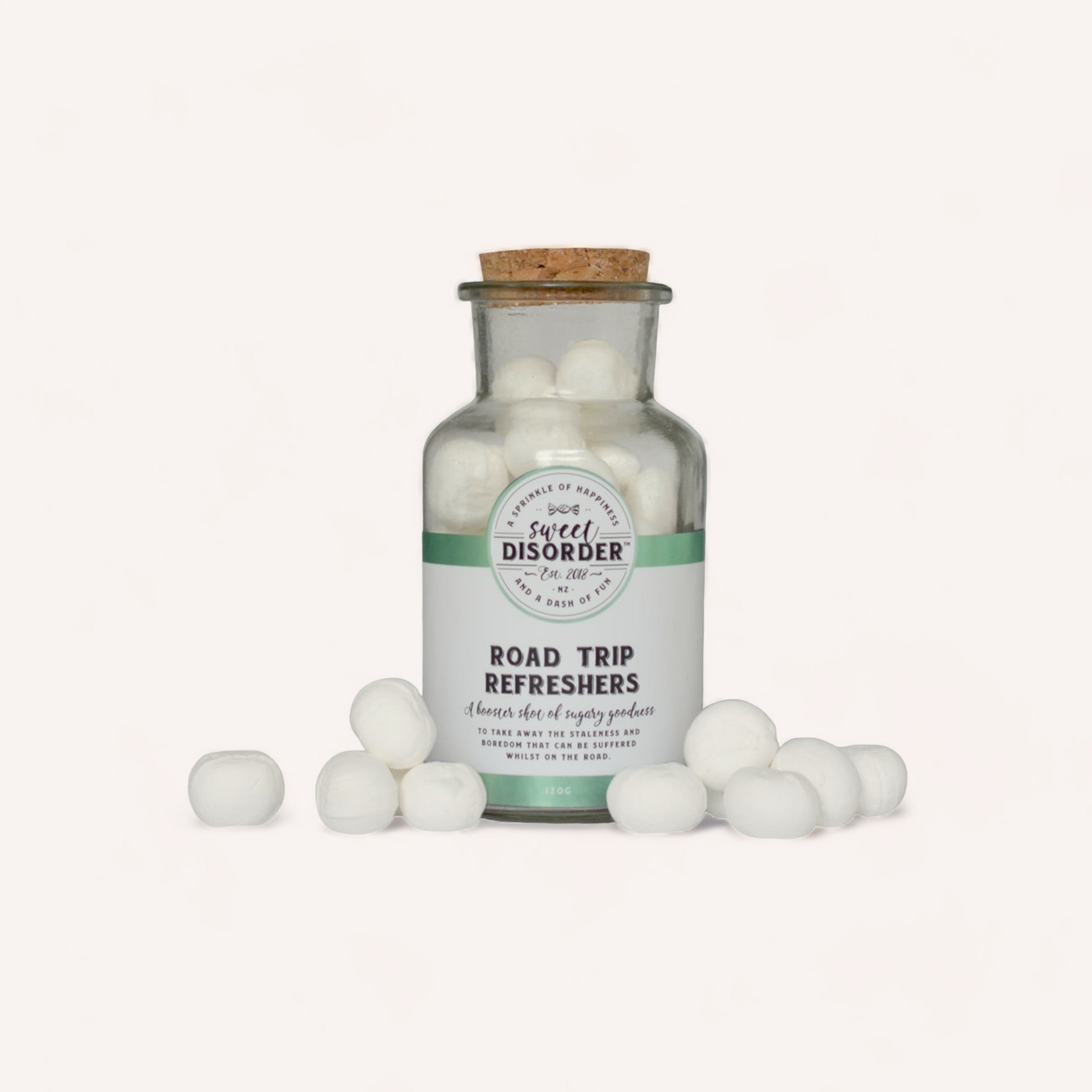 A glass jar with a cork stopper labeled "Road Trip Refreshers Lollies by Sweet Disorder," surrounded by white spherical contents scattered around it on a white background.
