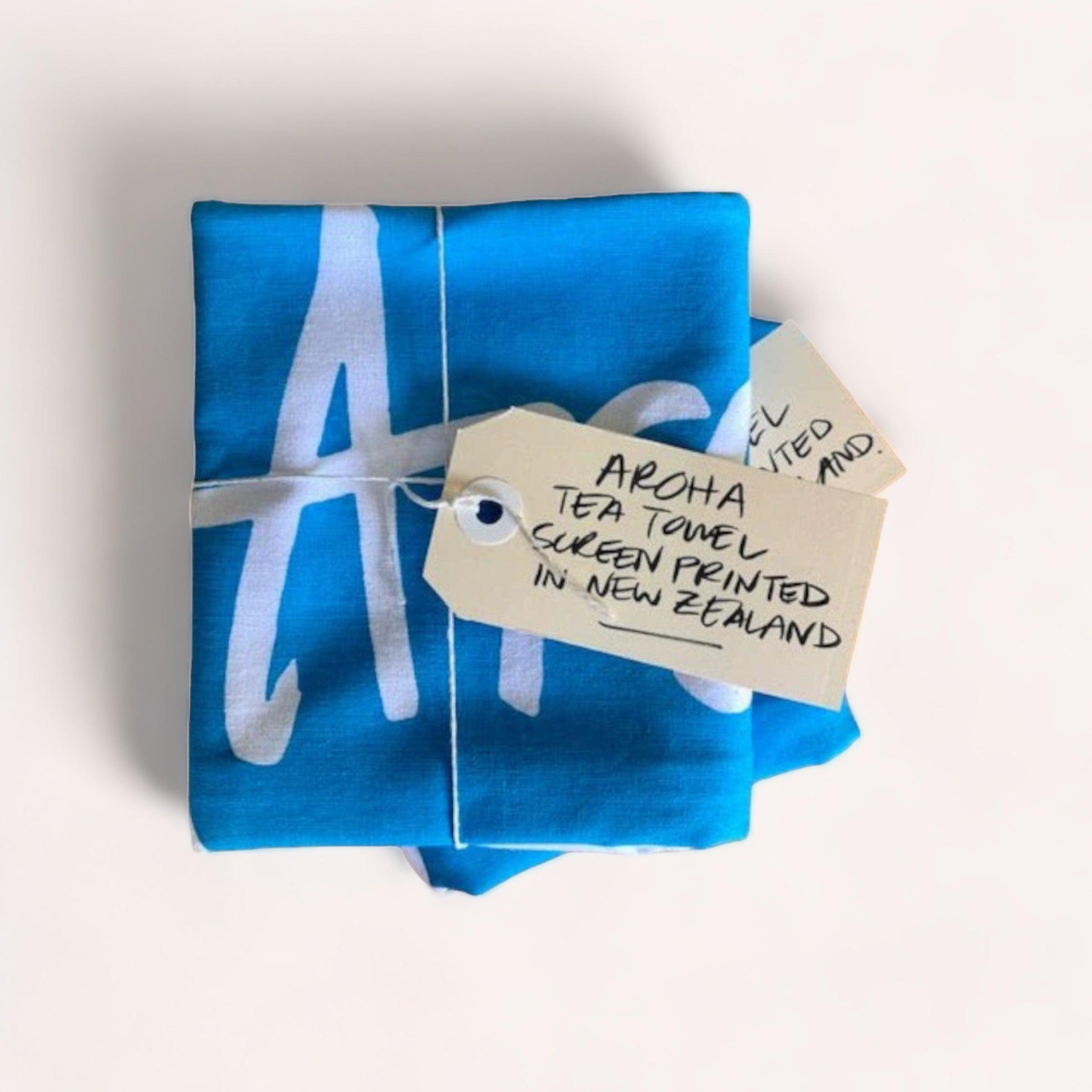 Folded blue fabric with white ‘a’ print and a tag indicating it is a 100% cotton, screenprinted Aroha Tea Towel from Tuesday Print in New Zealand.