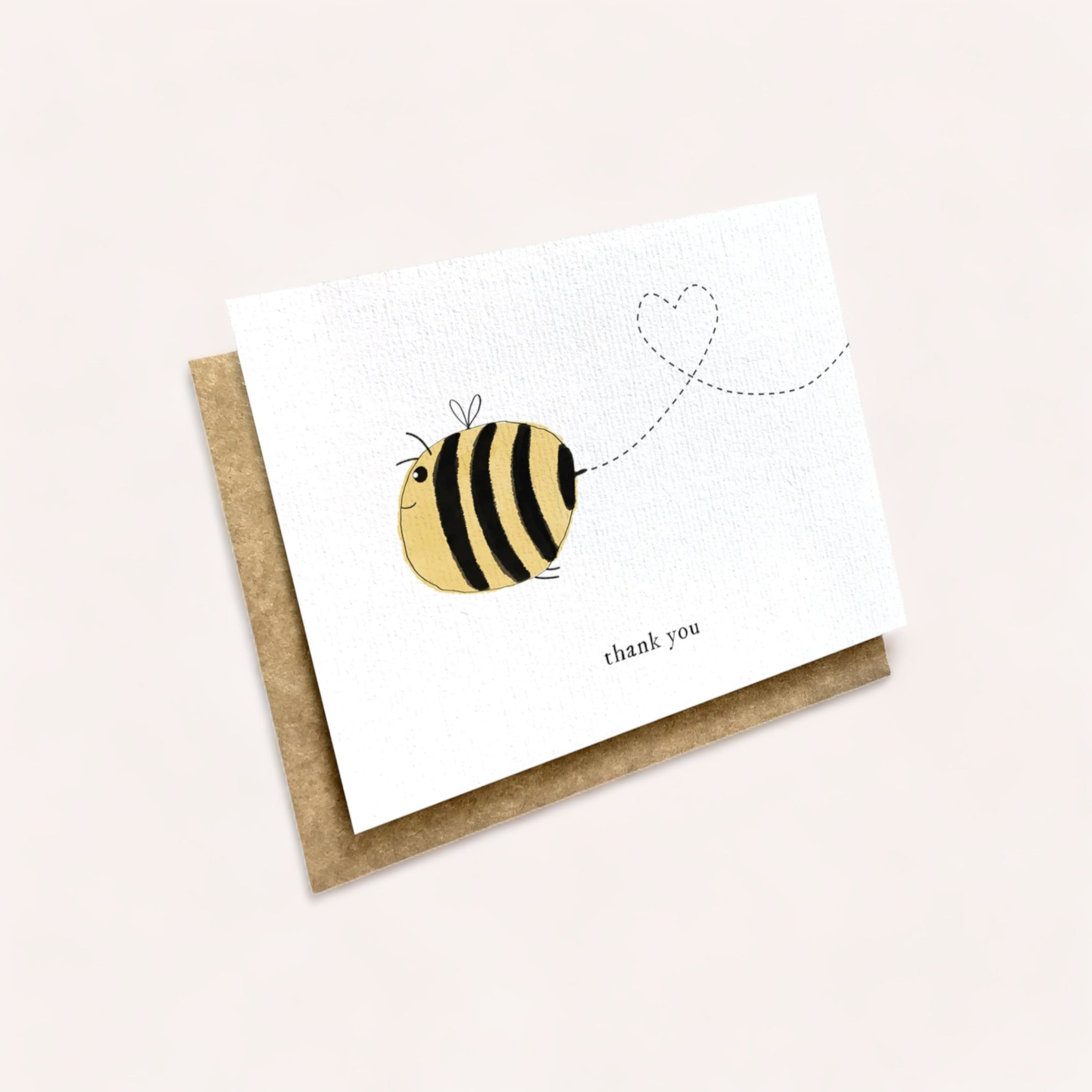 A Bee Thank You Card featuring a whimsical illustration of a bee with a heart-shaped flight trail on a plain background, accompanied by a kraft paper envelope, made by Ink Bomb.