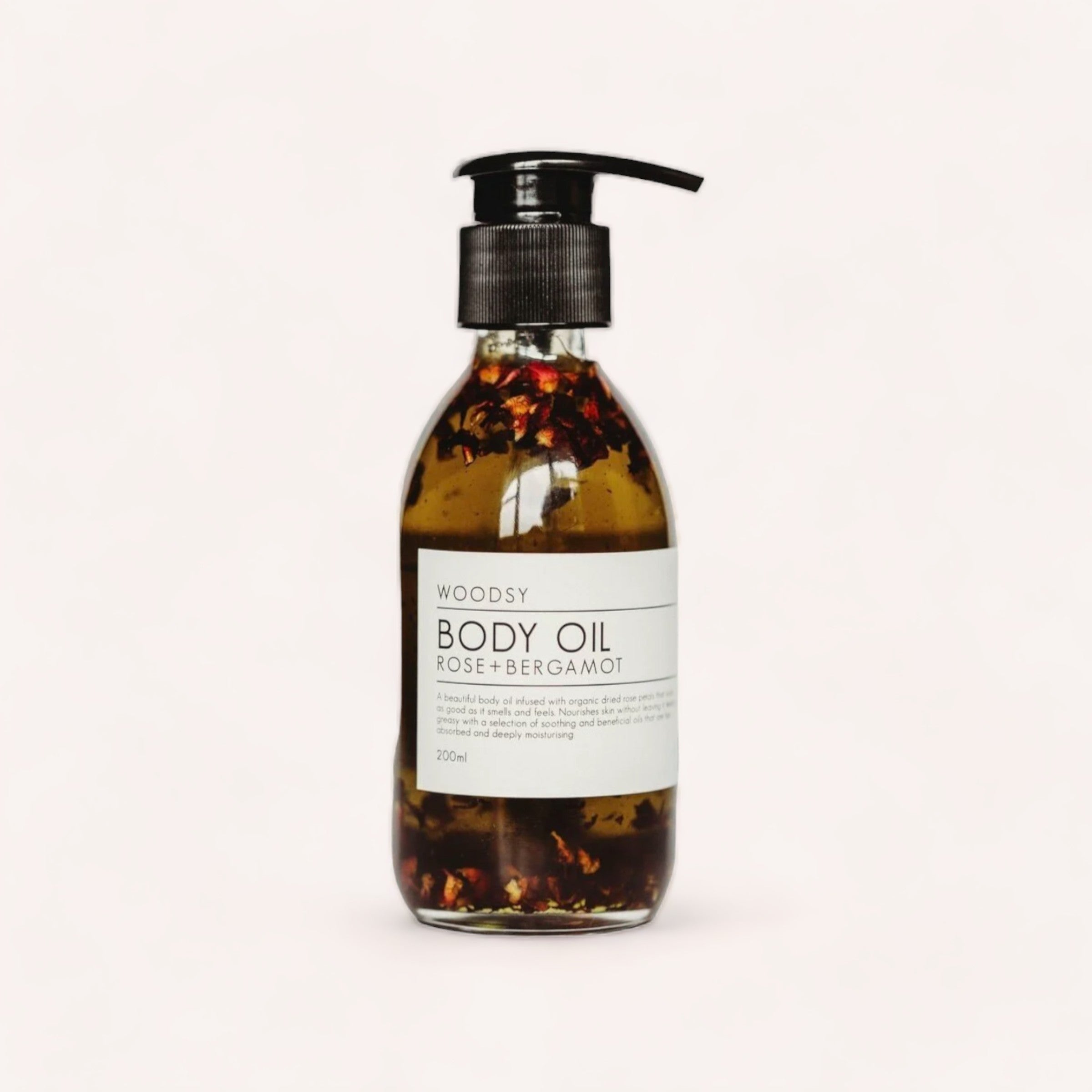 A clear bottle of Body Oil - Rose & Bergamot by Woodsy Botanics, with visible organic rose petals and leaves suspended in the oil, against a plain white background.