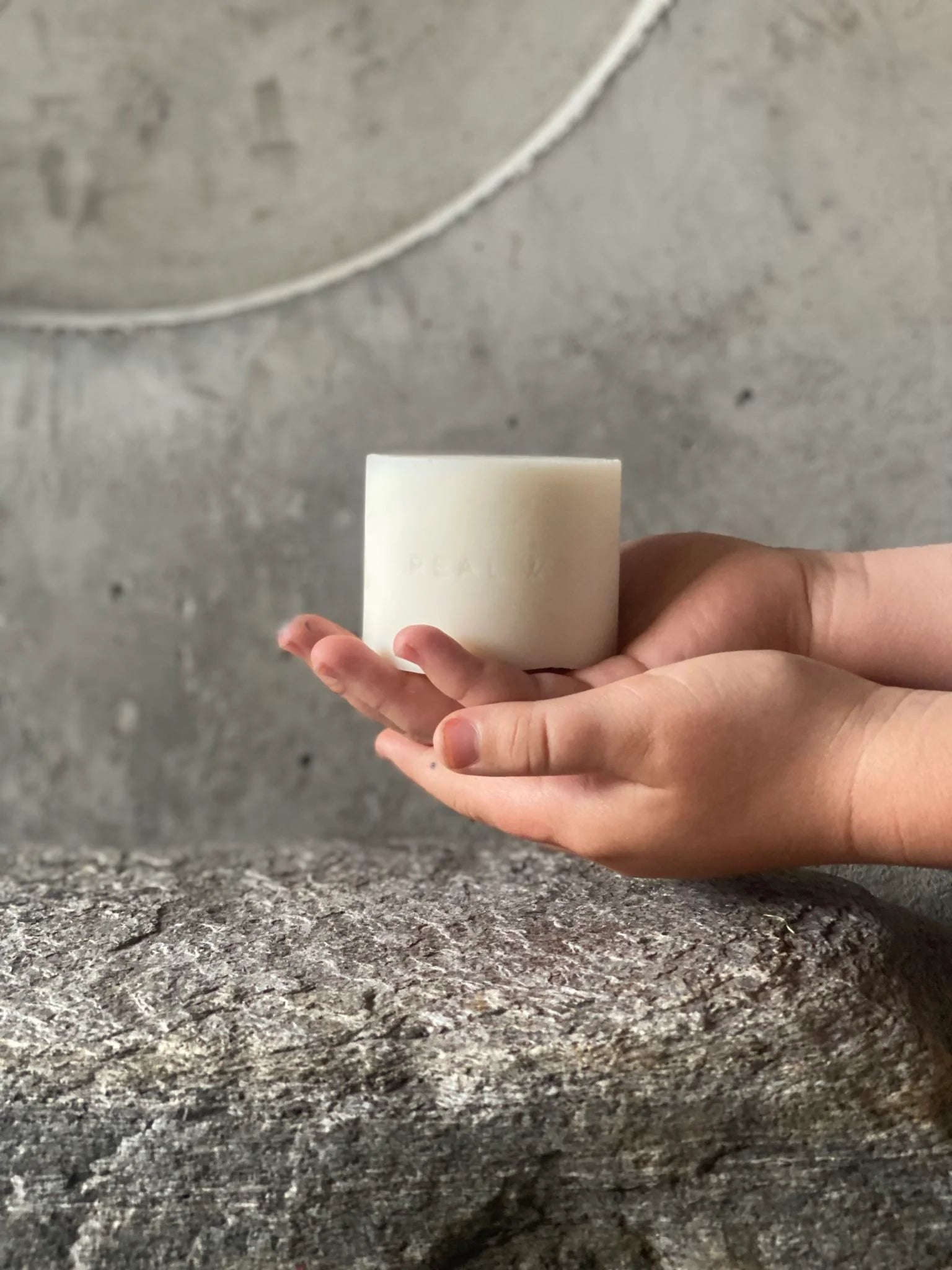 A child's gentle hands cradling a Soap Bar - Mamaku & Lavender by Real World, scented with lavender essential oils, set against a textured gray background.