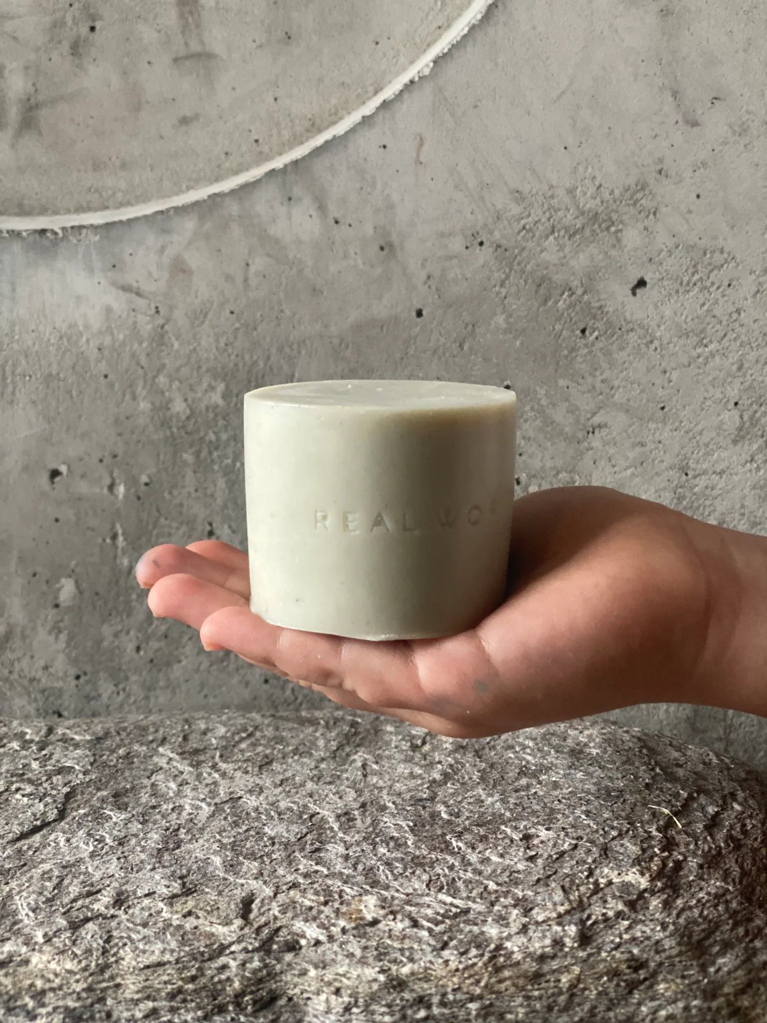 A gently held Soap Bar - Kawakawa, Clay & Palmarosa by Real World with the inscription "real love" against a concrete backdrop, symbolizing simplicity, serenity, and restorative cleansing.