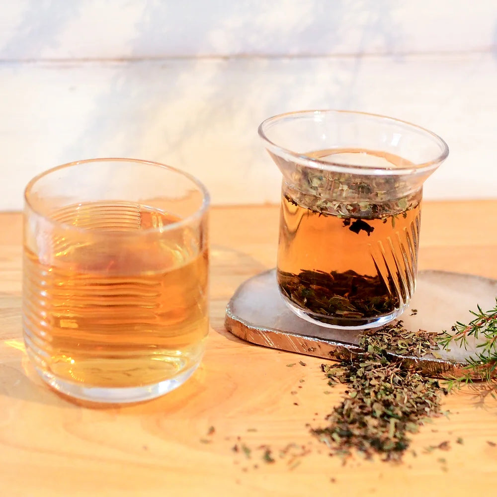 Two glasses of amber liquid, one with loose-leaf Summer Meadow 50g herbs infusing, on a wooden surface with scattered dried herbs and a sprig of thyme, offer an energy boosting refreshment by The Tea Thief.