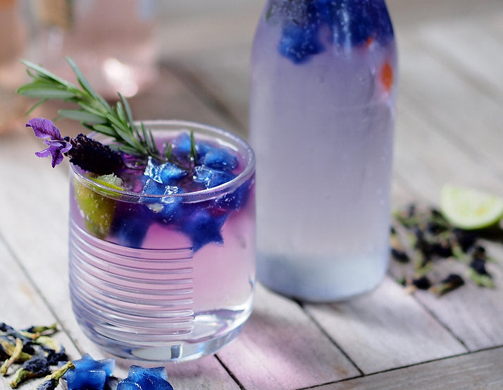 A refreshing glass of Tickety Brew 50g infused water adorned with vibrant edible lavender and a sprig of rosemary, accompanied by a frosted bottle and a slice of lime, set on a rustic wooden surface by The Tea Thief.