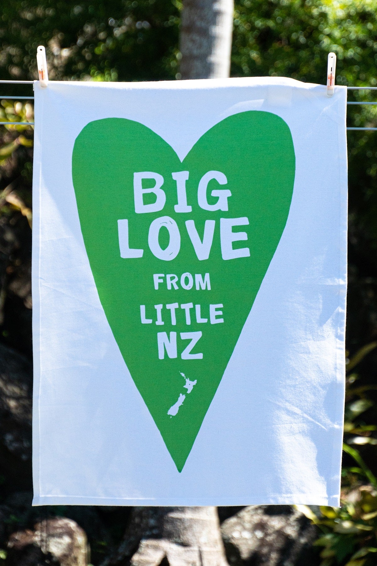 A vibrant, 100% cotton Big Love from little NZ Tea Towel by Tuesday Print with a large green heart and the message "big love from little nz" screenprinted on it, hanging on a clothesline, symbolizing warmth and affection.