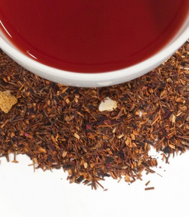 A close-up view of African Autumn Tea Tagalong by Harney & Sons in a white cup surrounded by loose dried tea leaves and bits of herbs against a white background.