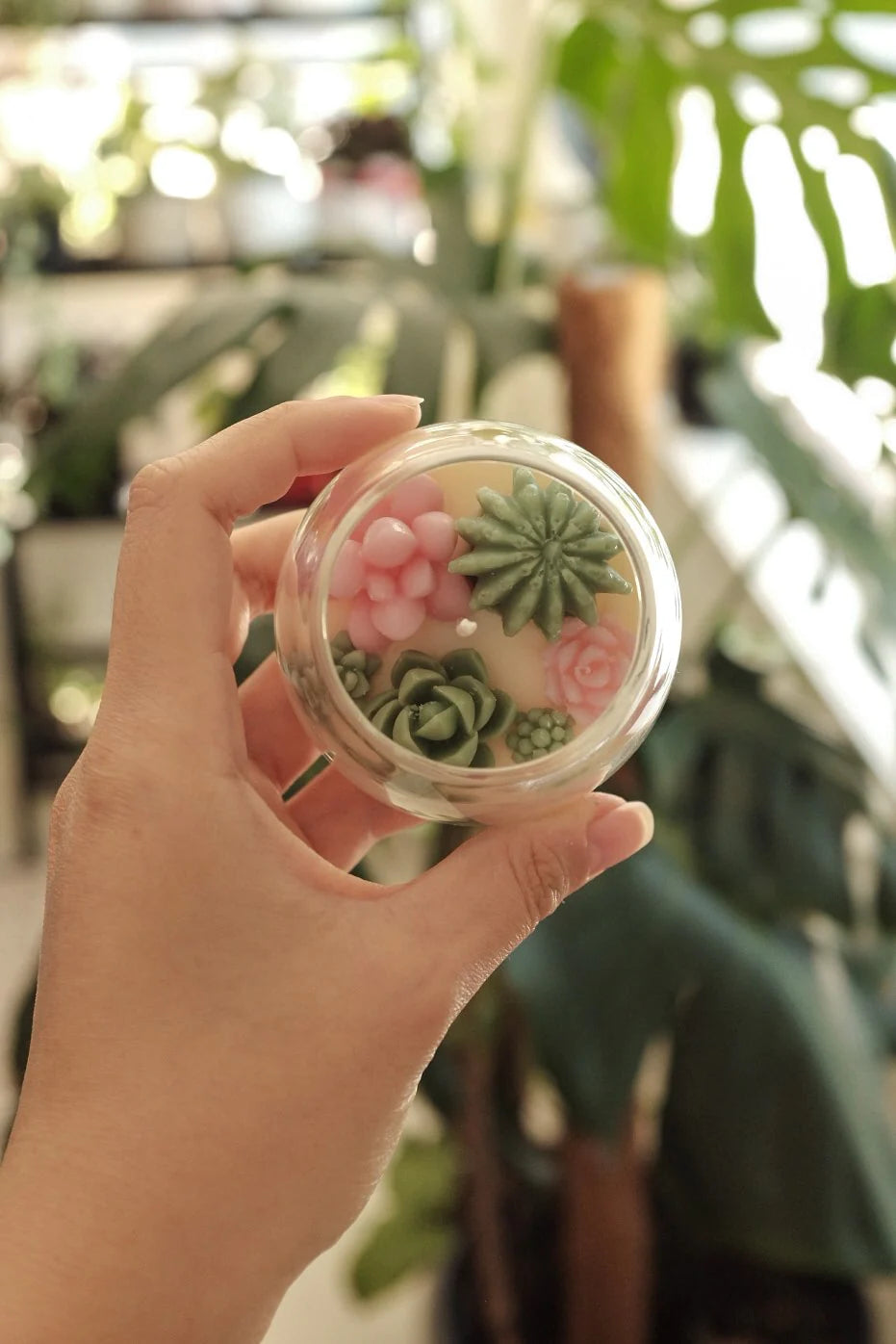 A person holding a small, transparent spherical container displaying a collection of delicate, lifelike succulents and flowers, including the Judean Desert Mini Terrarium Candle by Faith & Joy, against a backdrop of lush green plants.