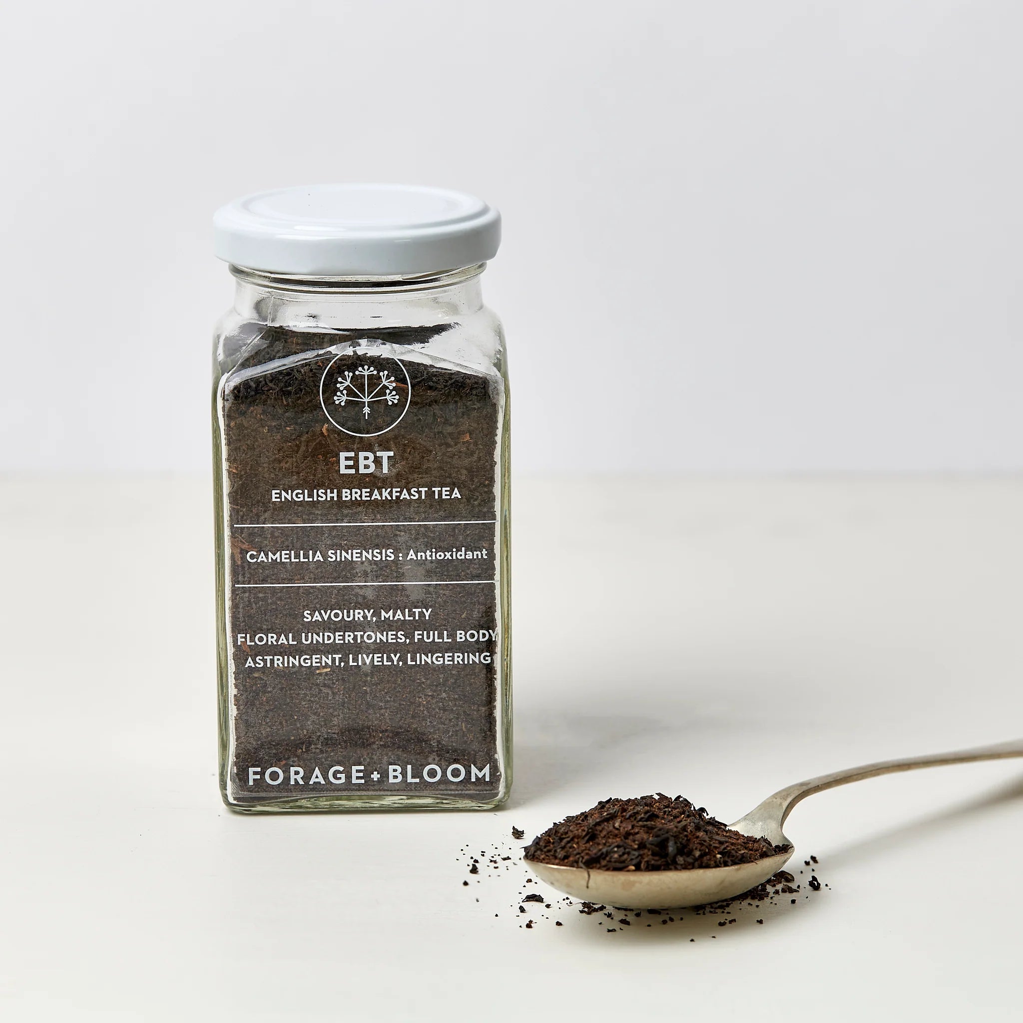 A jar of Tea for Two loose leaf English breakfast tea labeled with taste notes alongside a spoonful of loose tea leaves on a light background.