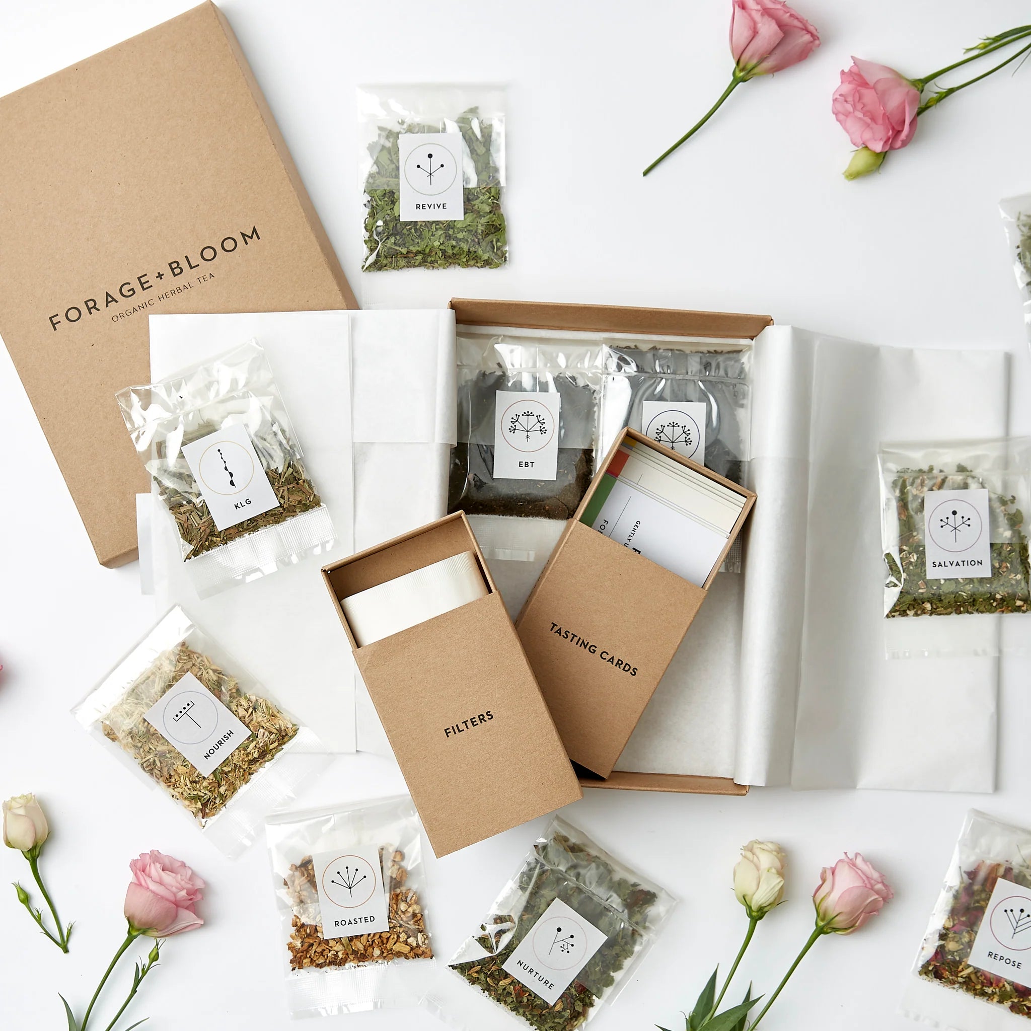 An assortment of elegantly packaged organic herbal tea blends, accompanied by the Tea Testing Box Set by Forage + Bloom and fresh flowers, thoughtfully presented in a gift box for a soothing and sophisticated tea experience.