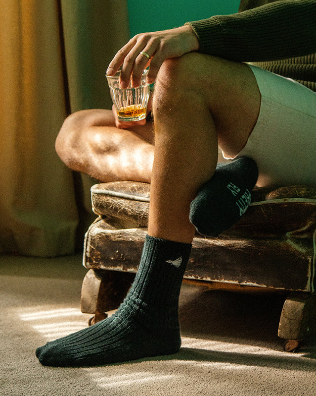 A moment of relaxation: a person unwinding in a cozy room with a drink in hand, bathed in warm sunlight, and clad in sustainably produced Marino Wool Socks from Pretty Fly.