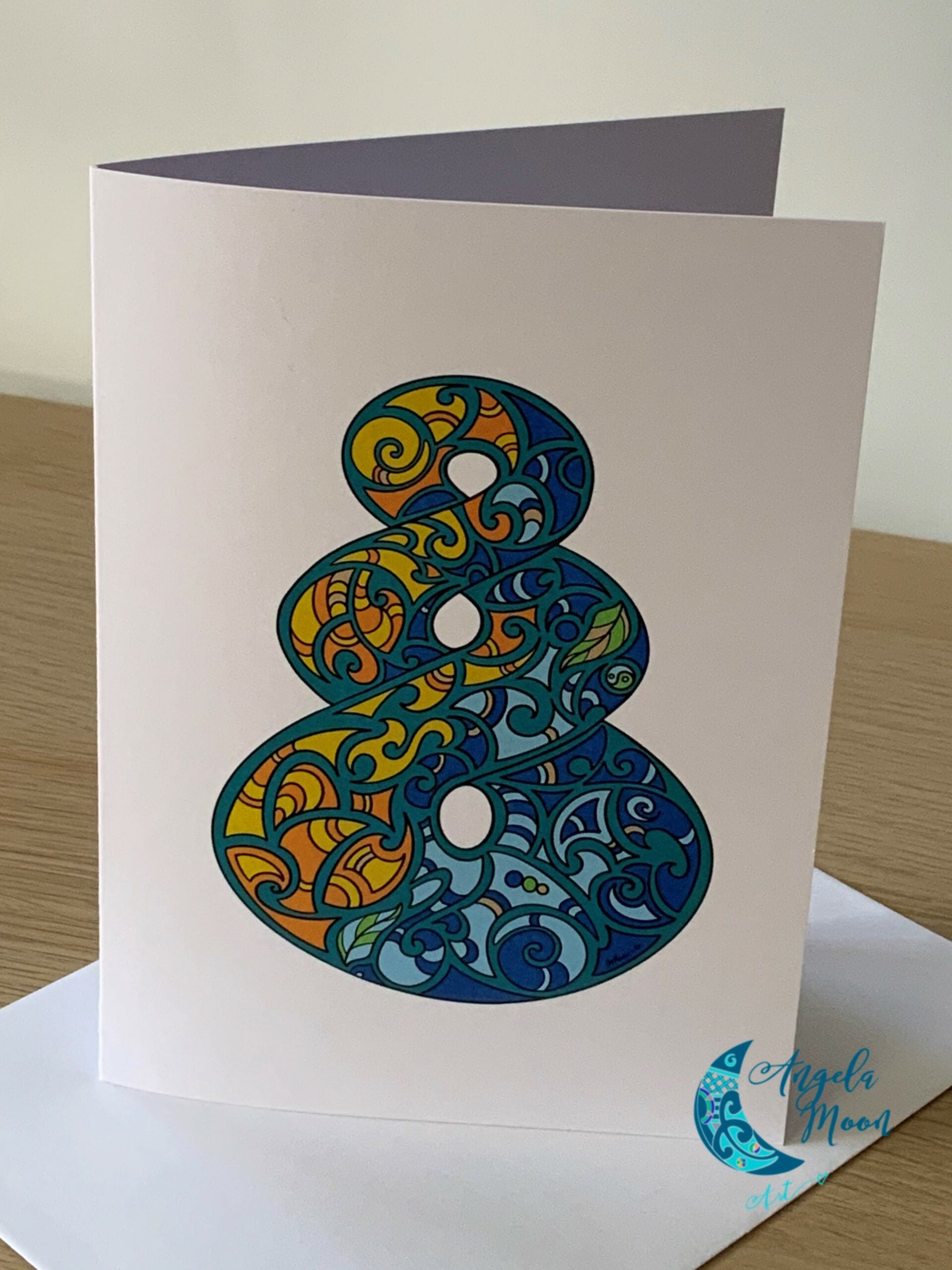 A colorful, artistic Pikorua Card from Angela Moon Art in New Zealand with intricate patterns displayed alongside its envelope on a wooden surface.