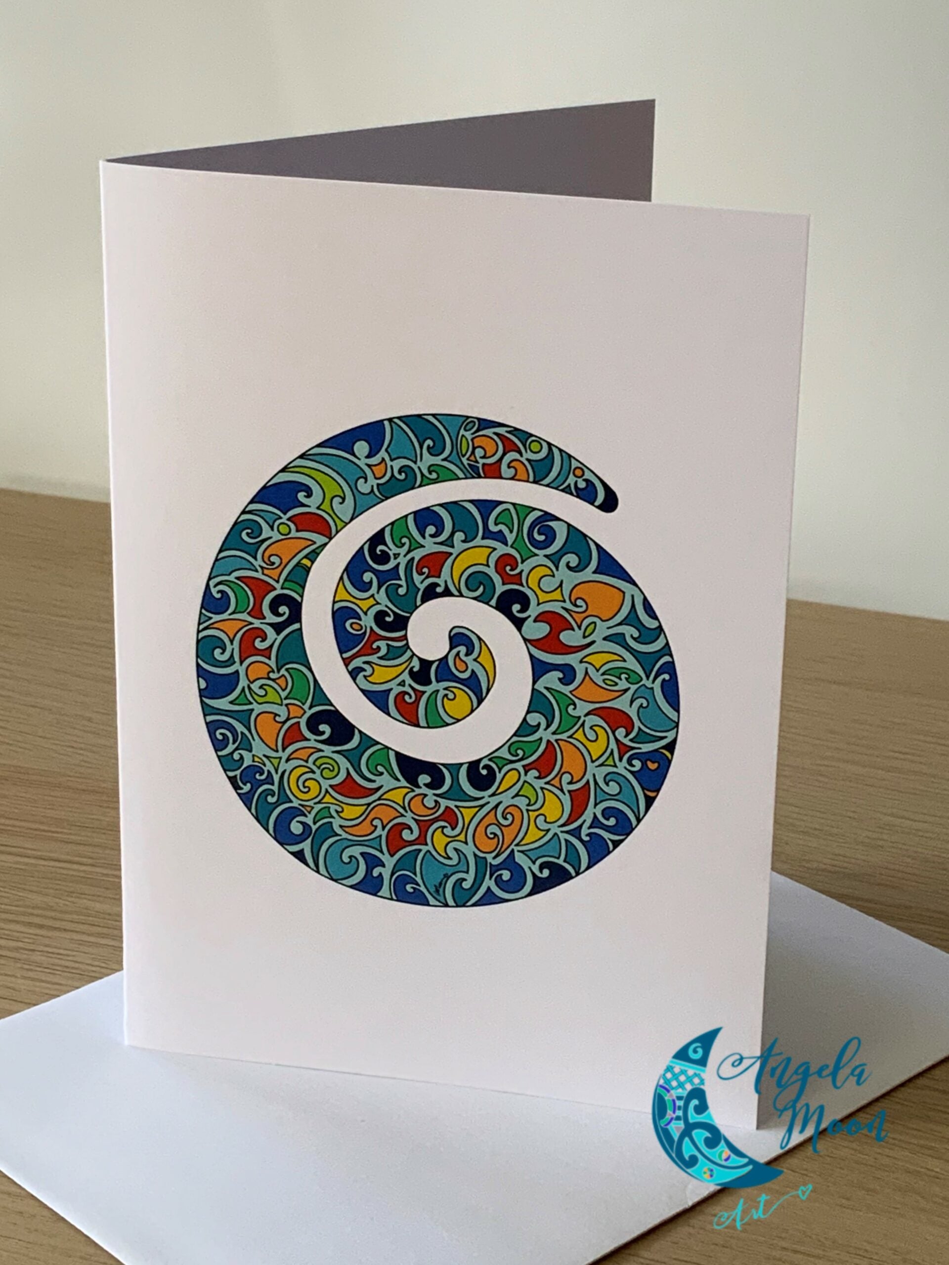 A colorful and intricately designed Angela Moon Art Koru Fern Frond Card with a spiral pattern resembling a paisley, displayed with its envelope on a wooden surface.