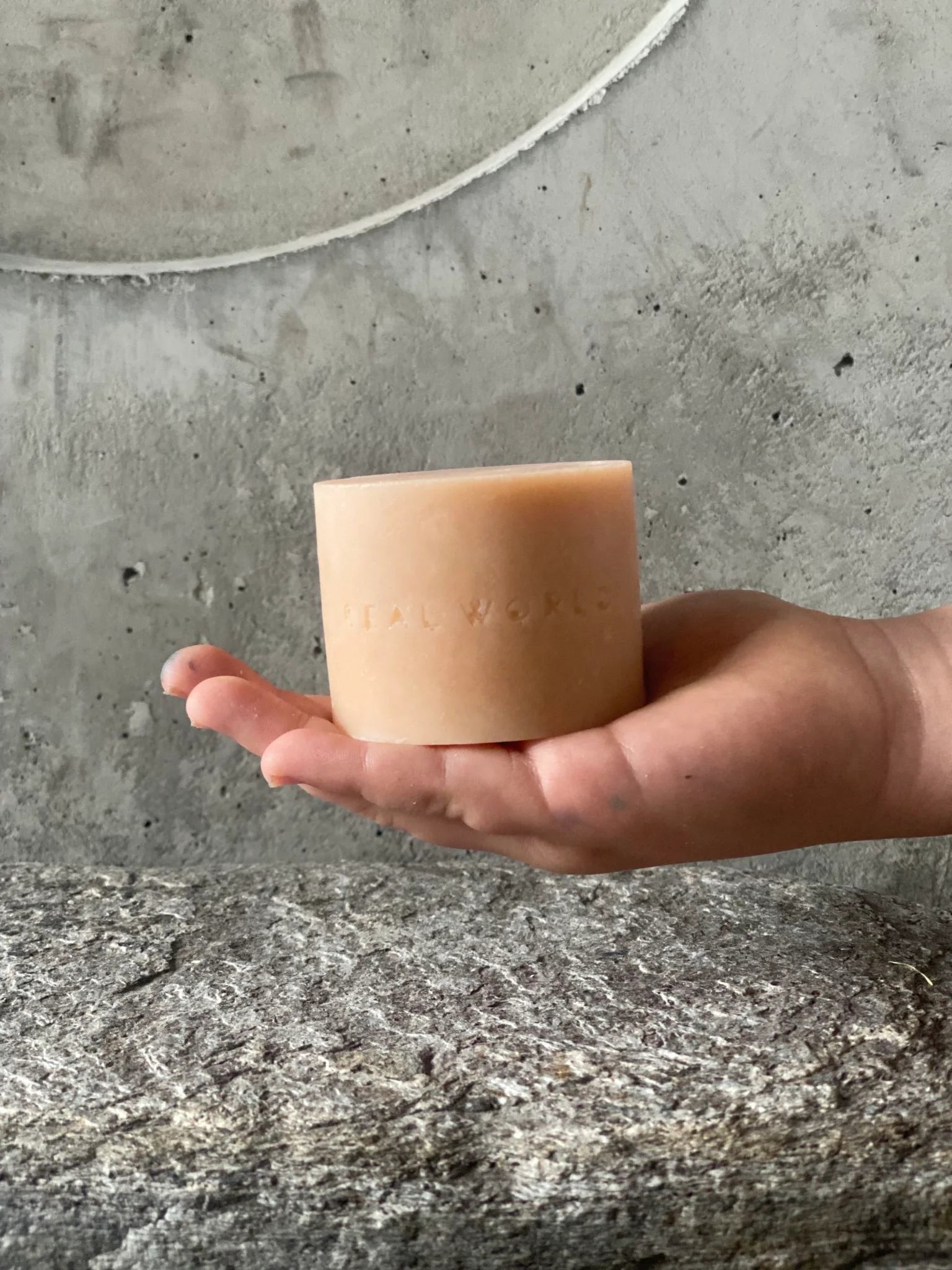 A hand gently holding a Soap Bar - Manuka & Rose Geranium by Real World with the words "Italy Works" embossed on it, against a textured grey concrete background.