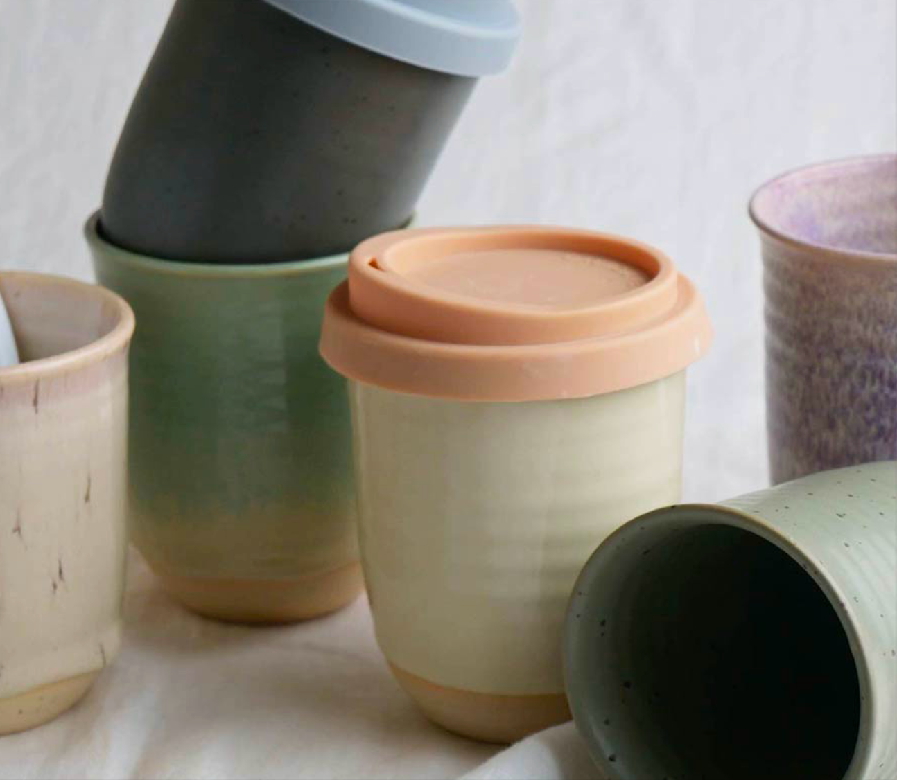 An assortment of Ceramic Keep Cups by Sam Mayell in various colors, some with lids, arranged casually on a pale surface.