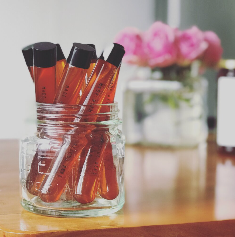 A charming arrangement: Honey Vial 38ml lip glosses, reminiscent of raw natural honey and proudly a Product of New Zealand, fashionably showcased in a clear glass jar, with a soft-focus bouquet of pink by Bees Up Top.