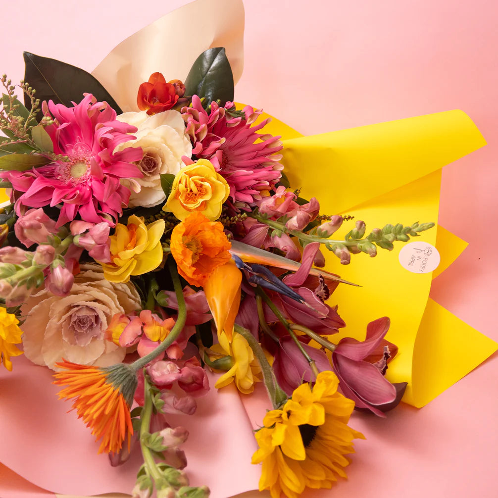 A vibrant bouquet of Doreen Bright & Vibrant flowers in shades of pink, yellow, and orange from our bespoke floral collection, elegantly wrapped in yellow paper against a soft pink background. Brand Name: Poppy in April