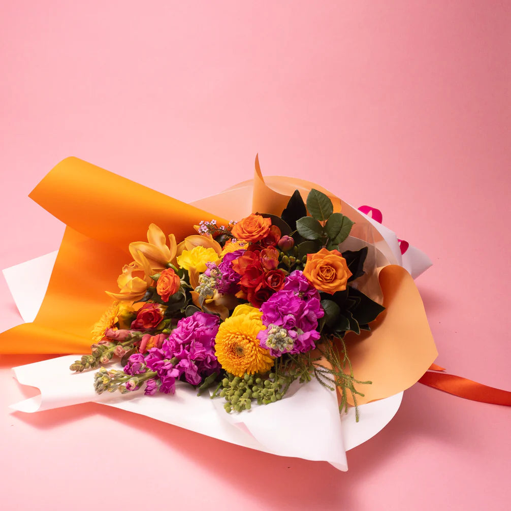 A vibrant bouquet of Doreen Bright & Vibrant floral arrangements wrapped in orange and white paper against a pink background by Poppy in April.