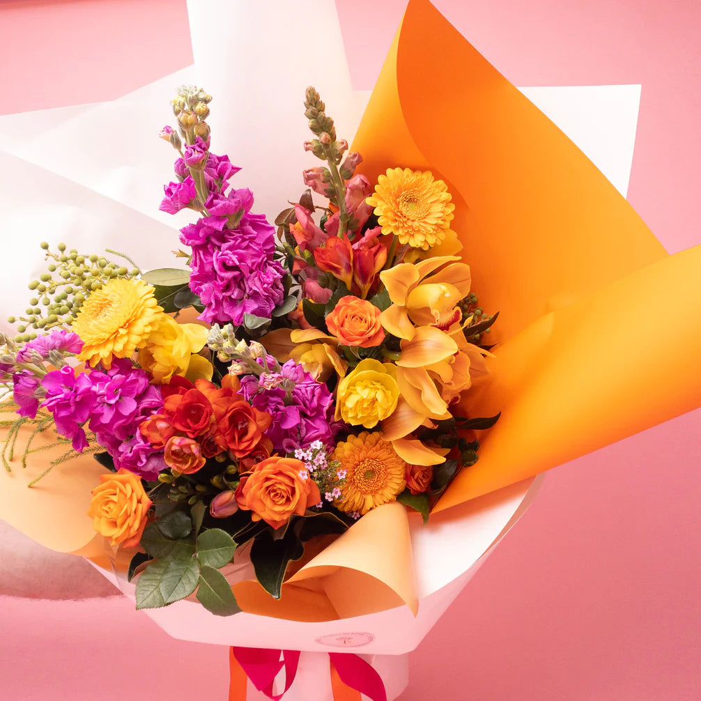 A vibrant bespoke floral bouquet with a mix of purple, yellow, orange, and pink blooms wrapped in a contrasting orange and white paper on a pink background by Poppy in April's Doreen Bright & Vibrant.