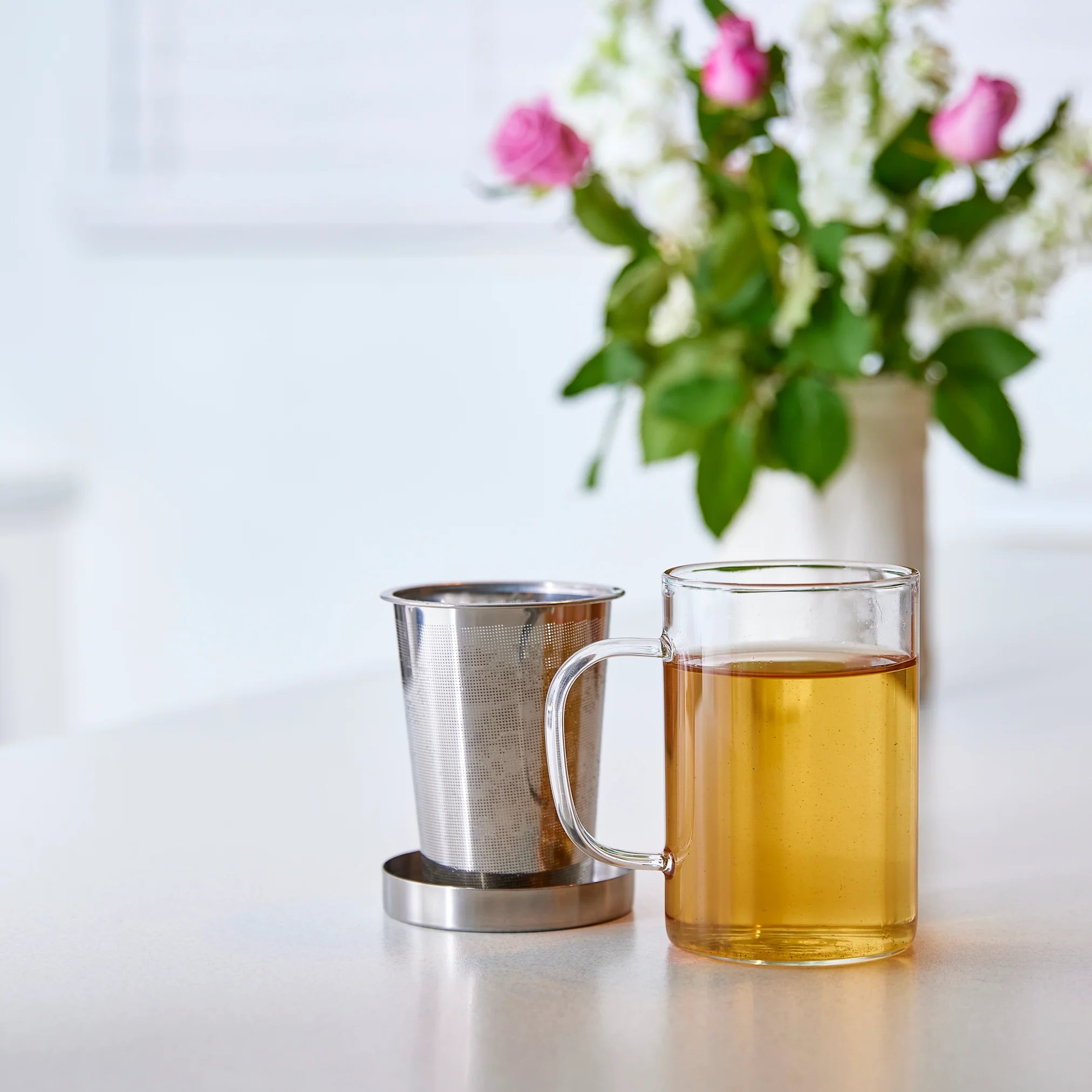 A Forage + Bloom heat-resistant glass tea cup filled with amber tea beside a Forage + Bloom stainless steel infuser for loose leaf brewing on a white surface, with a vase of roses in the soft-focus background.
