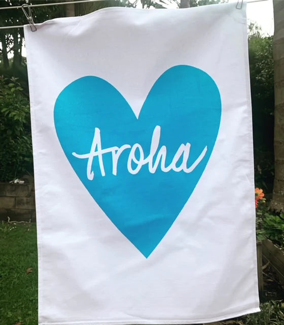 A white flag with a large turquoise heart and the word "aroha" screenprinted across it, displayed in a garden setting - Aroha Tea Towel by Tuesday Print.