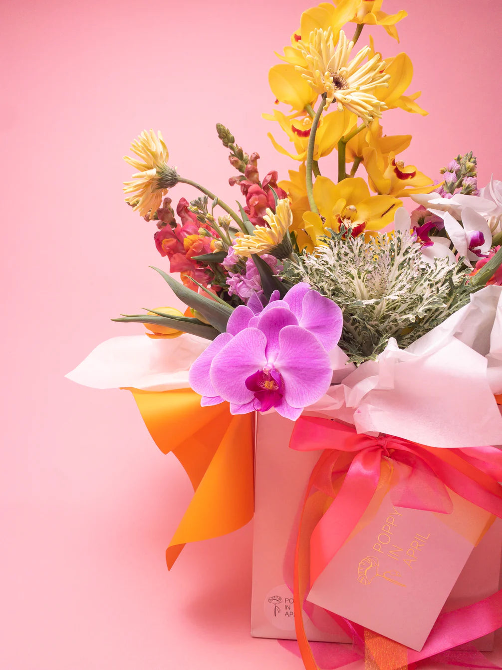 A vibrant bouquet of yellow and pink seasonal blooms with a prominent orchid, elegantly wrapped and adorned with a pink ribbon and a gift tag, set against a pink background. The Annie bouquet from Poppy in April.