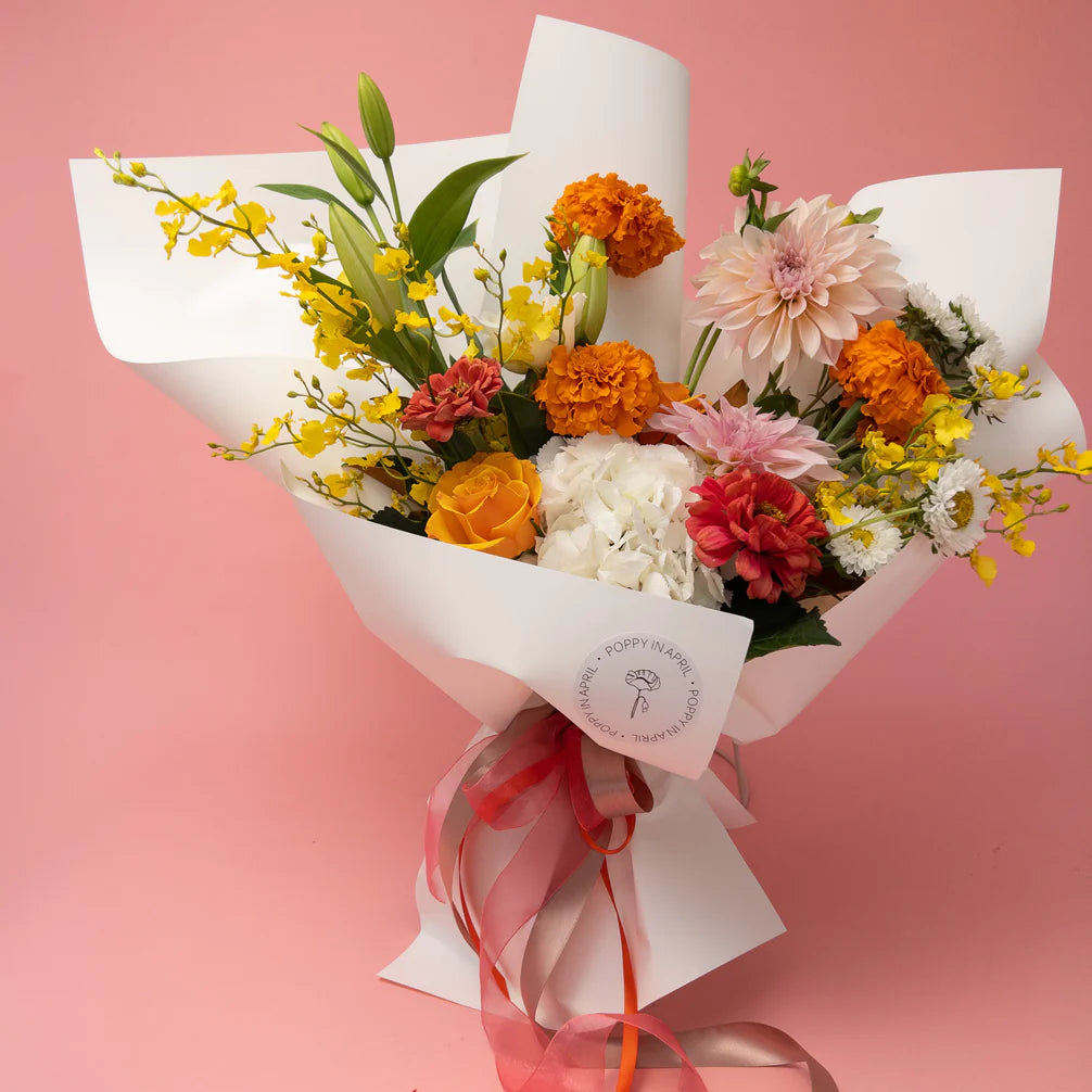 A vibrant bouquet of Poppy in April's Florist Choice mixed seasonal florals wrapped in white paper with a ribbon and a bespoke gift message against a pink background.