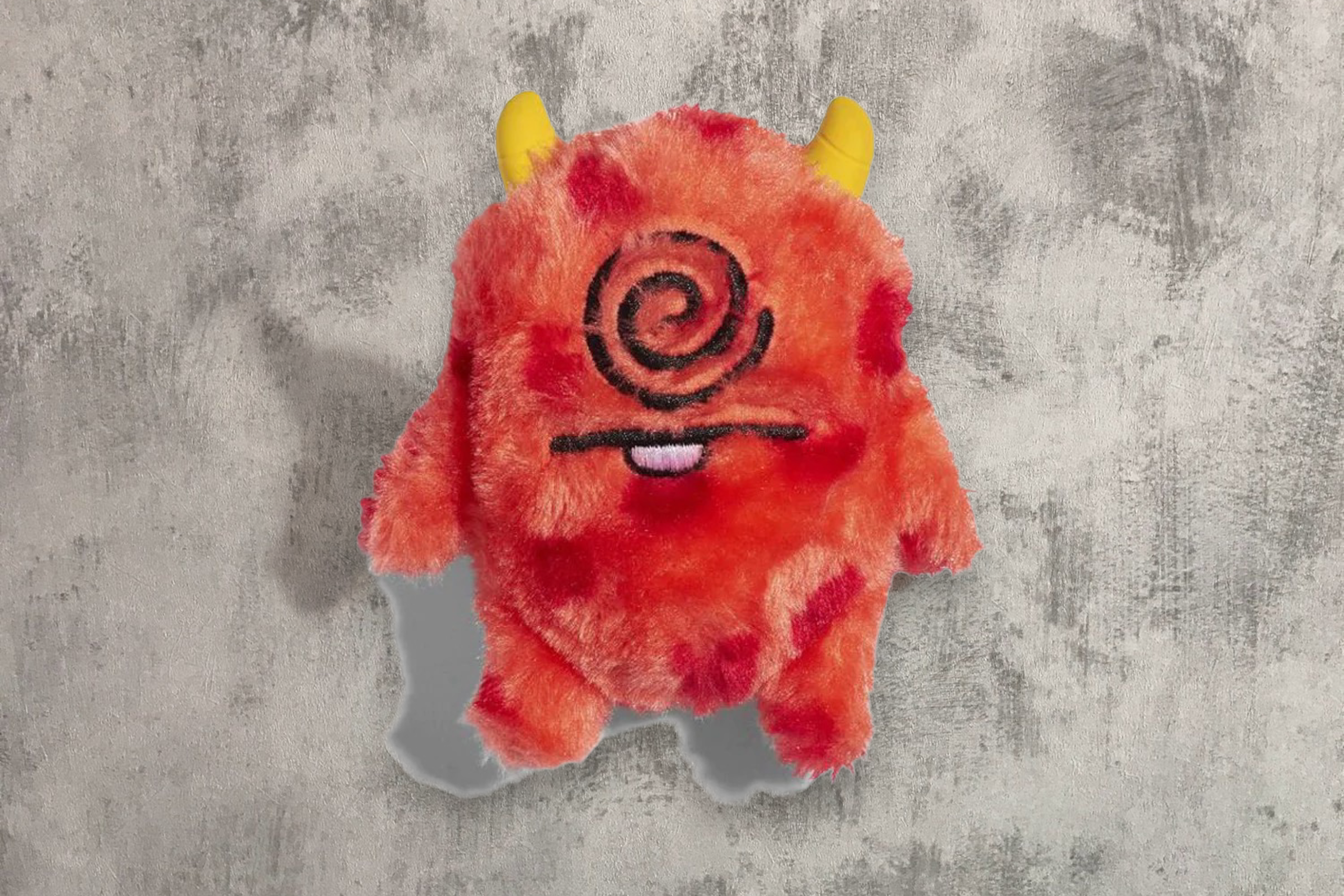 A Cyclops Pet Toy by Zee.Dog plush toy with a whimsical red and orange fur design, featuring a Cyclops swirled eye and a playful expression, complete with little yellow horns, set against a textured grey background.