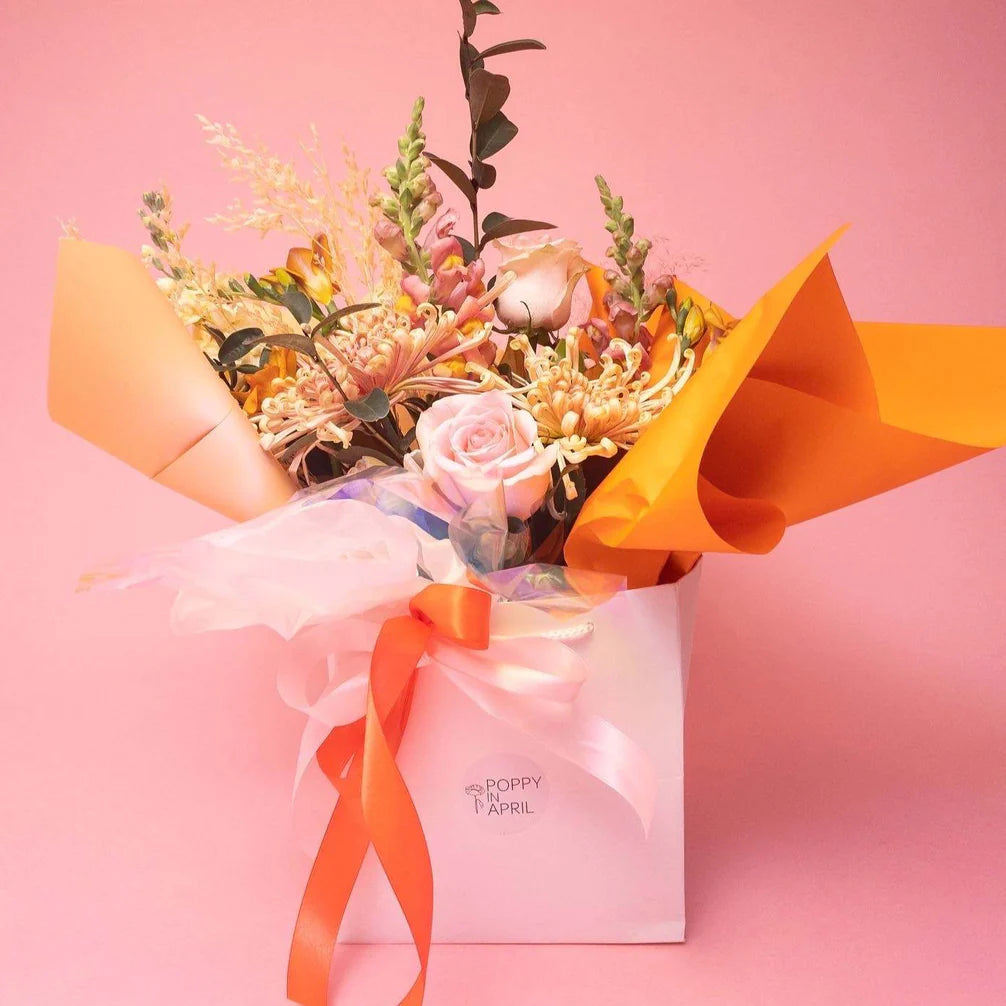 A vibrant bouquet of seasonal flowers with a mix of roses and assorted blooms, tastefully wrapped and presented in a stylish white bag with the text "Masie," set against a soft pink background by Poppy in April.