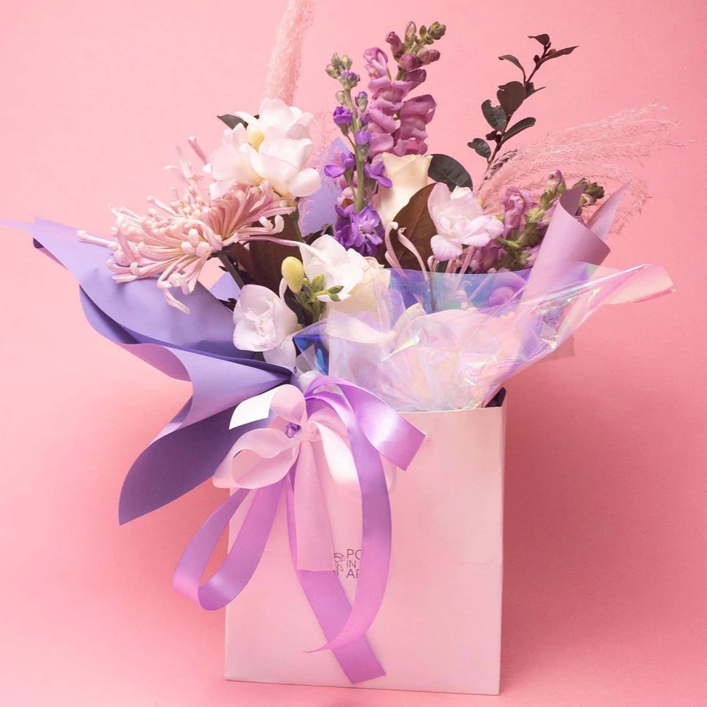 A charming bouquet of mixed seasonal flowers in shades of pink, white, and purple, elegantly wrapped in blue paper and tied with a satin ribbon, presented in a stylish pink Poppy in April box.