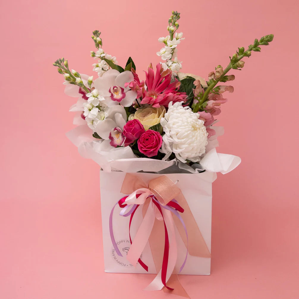 A charming bespoke floral arrangement featuring a blend of white and pink seasonal flowers with lush greenery, presented in a white gift box adorned with a pink ribbon, set against a soft pink background by Poppy in April's Masie.