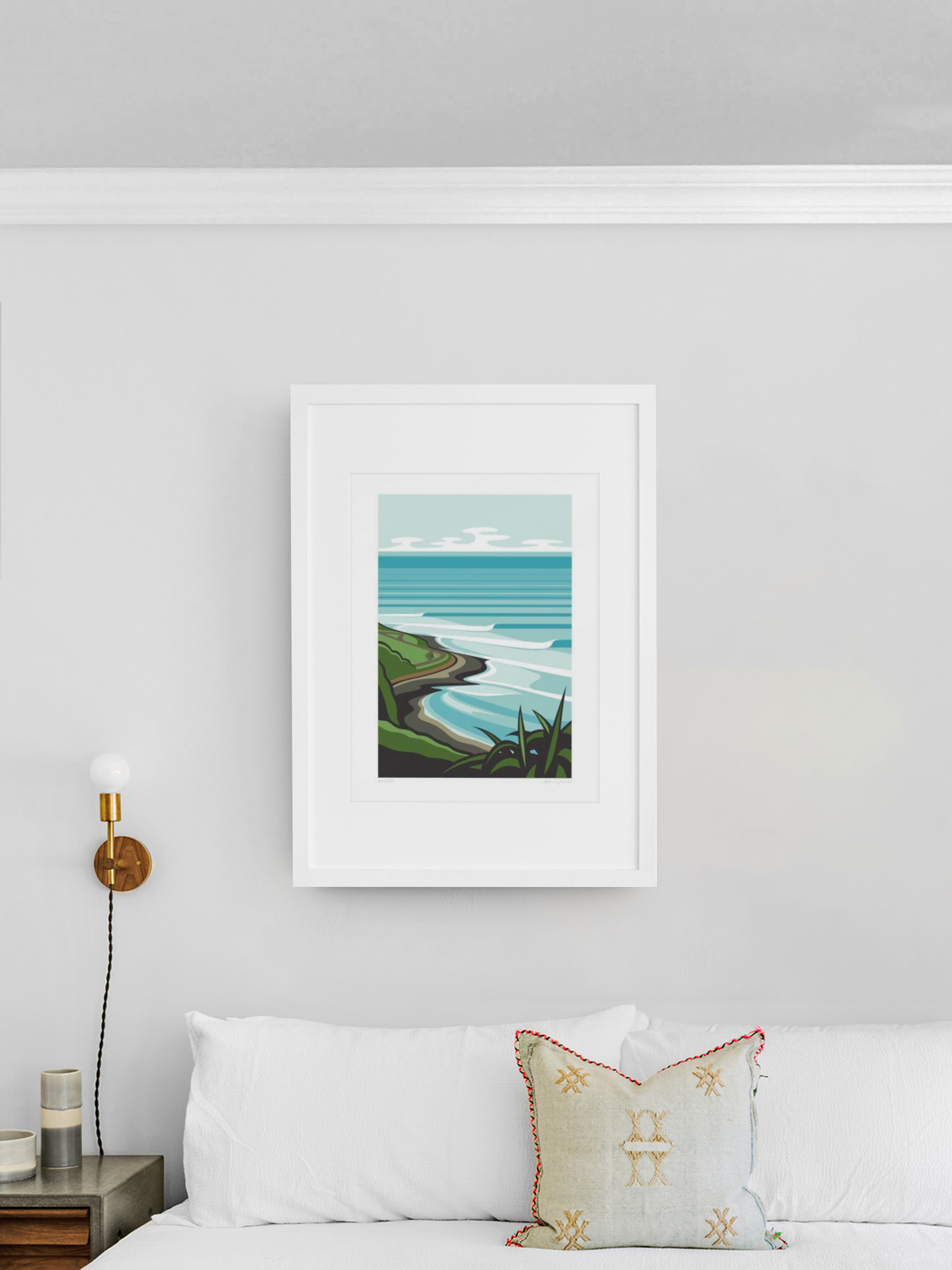 A sleek white-framed piece of art from the Scenic Series, depicting Raglan by Glenn Jones, hangs elegantly on a light gray wall above a cozy corner of a white sofa, adorned.