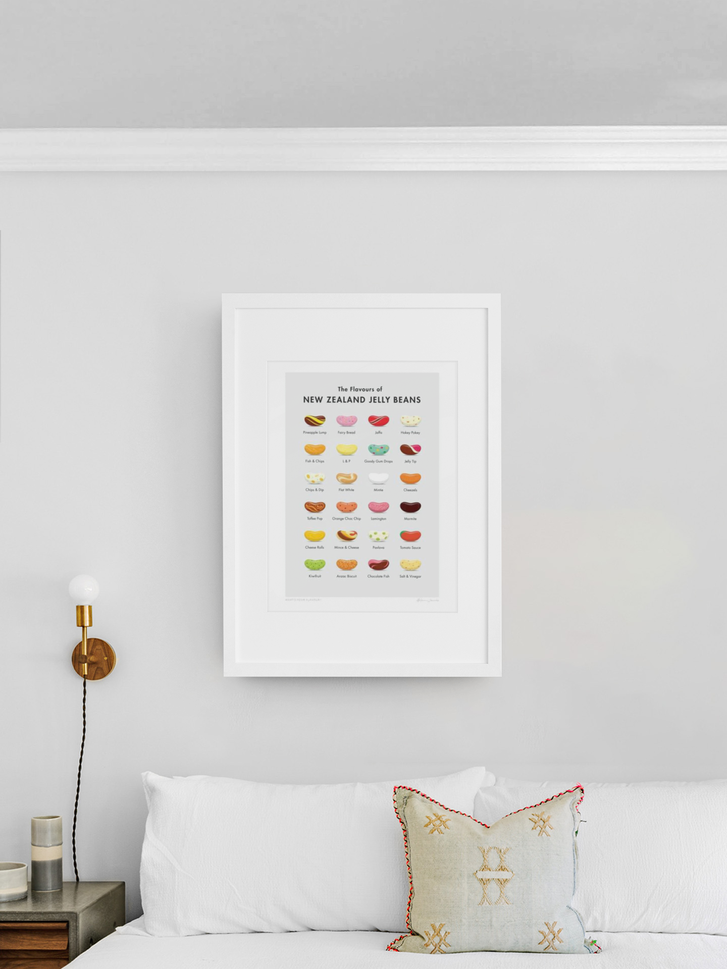A clean and minimalistic room featuring a framed poster of "What's Your Flavour? by Glenn Jones" with colorful illustrations hung on a plain white wall above a cozy white sofa with an ornate pillow. The poster highlights Glenn Jones.