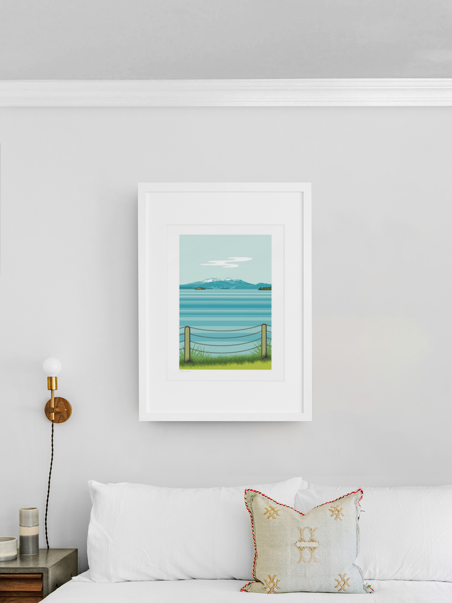 A serene Lake Taupo painting from Glenn Jones' Scenic Series in a white frame adorns a clean and modern living room wall, above a white couch with an elegantly patterned cushion.