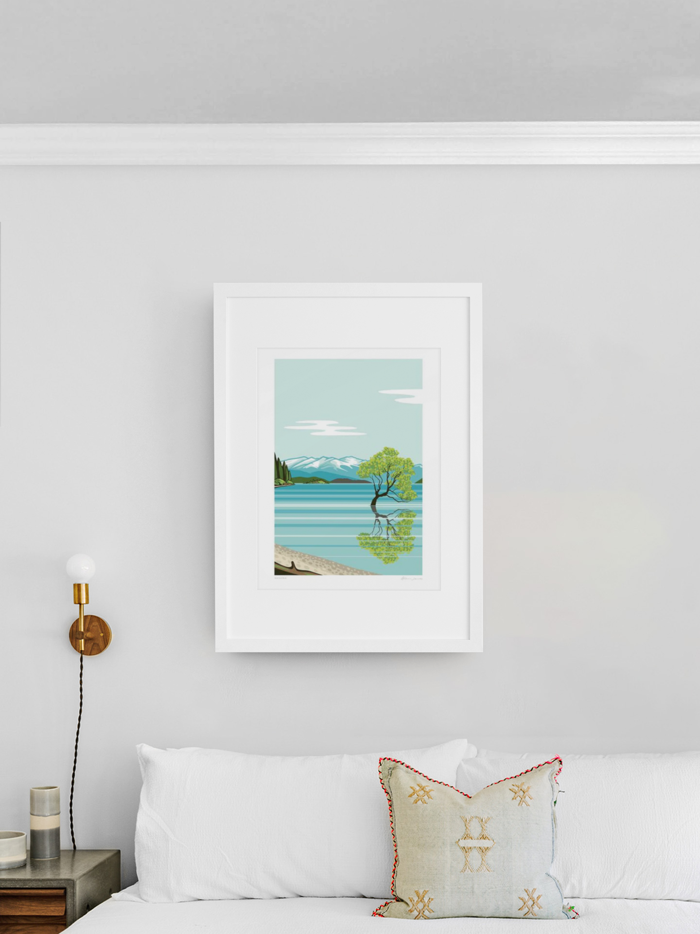 A serene landscape painting from the Wanaka by Glenn Jones series featuring the Wanaka Tree and a lake is elegantly framed and displayed on a clean, white wall above a cozy and inviting bed with plush pillows.