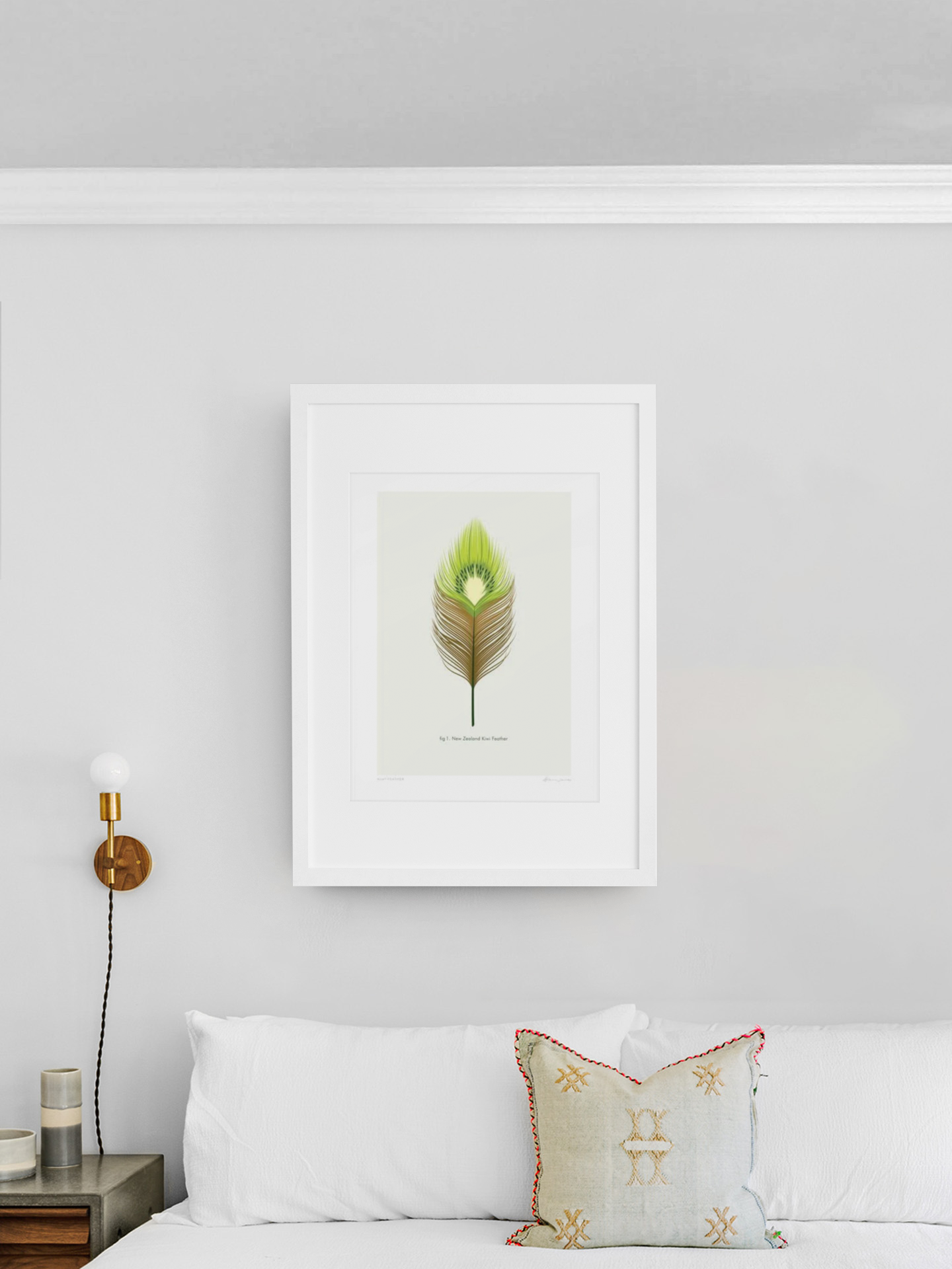 A single, vibrant green Kiwi Feather by Glenn Jones, a product of New Zealand, framed and displayed on a clean white wall above a cozy white sofa adorned with a decorative pillow, accompanied by a stylish wall-mounted.