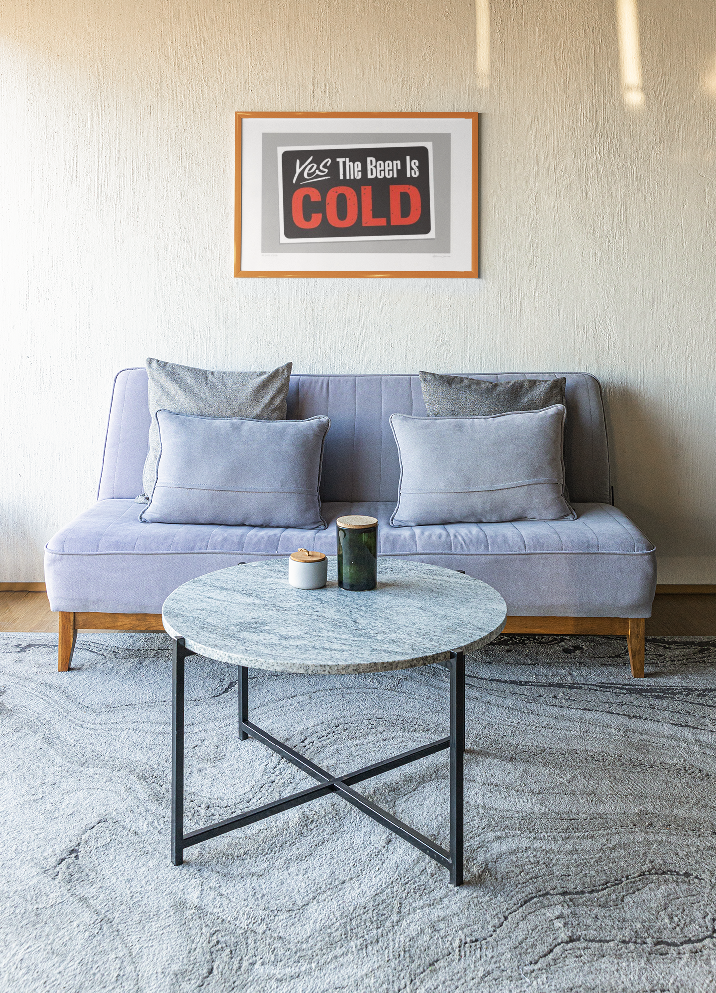 A cozy living room setup with a modern gray sofa, Beer is Cold by Glenn Jones, decorated with matching pillows, a round marble-top coffee table with a candle and a tumbler on it, set on a textured gray rug.