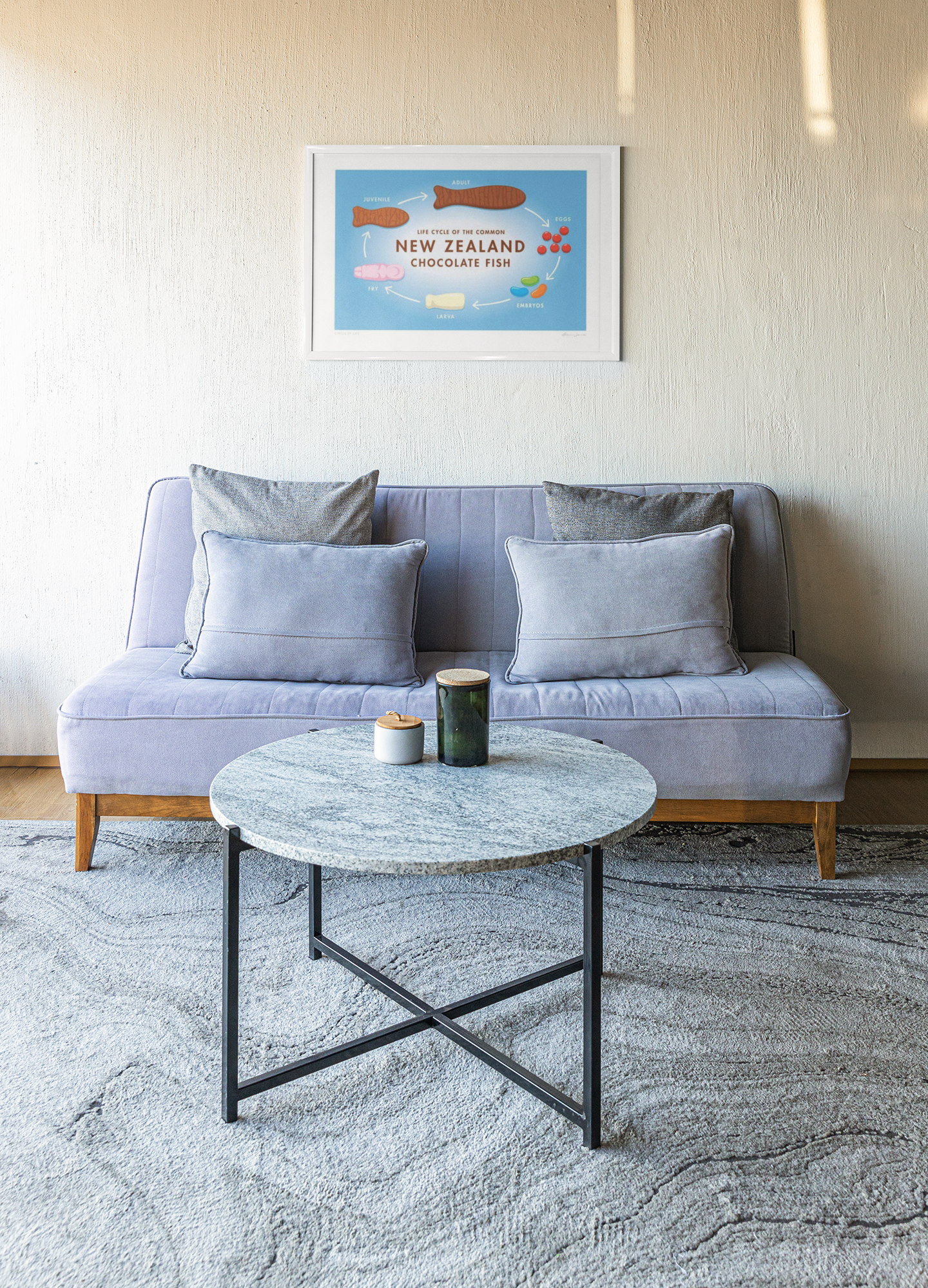 A cozy living area featuring a modern gray sofa adorned with cushions, a round coffee table with a candle and a cup, all set on a textured rug, with a framed Glenn Jones Circle of Life poster.