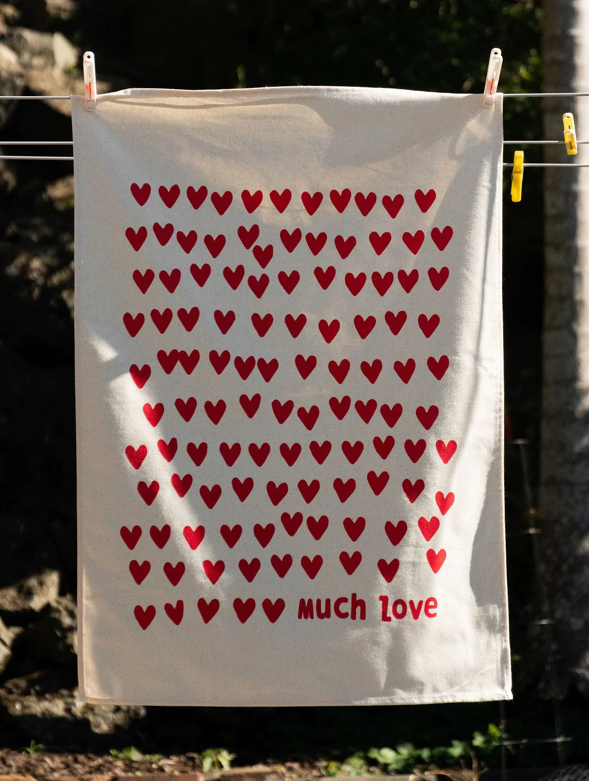A Much Love Tea Towel adorned with multiple red hearts in a grid pattern and the words "much love" at the bottom, hanging on a clothesline outdoors by Tuesday Print.