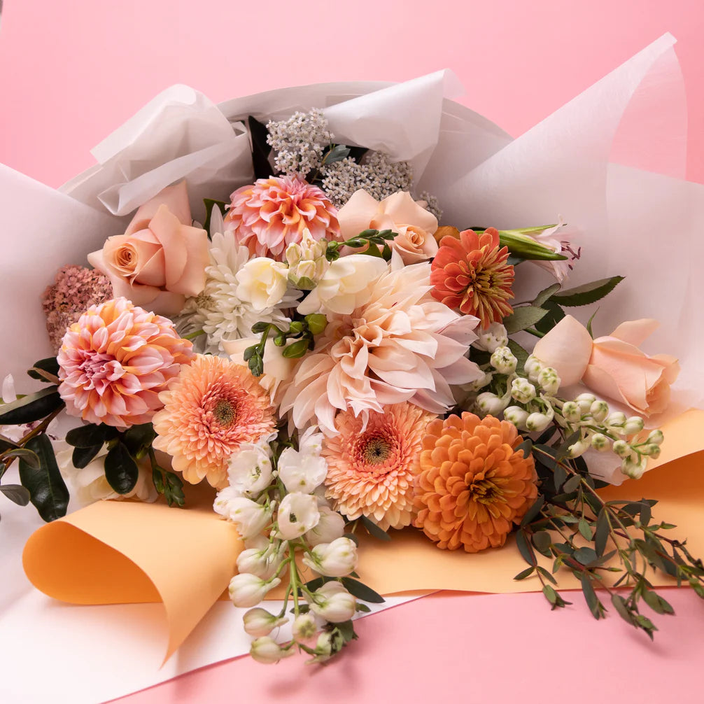 A vibrant bouquet of Doreen Pretty & Pastel flowers from Poppy in April's bespoke floral collections, wrapped in white and yellow paper on a pink background.