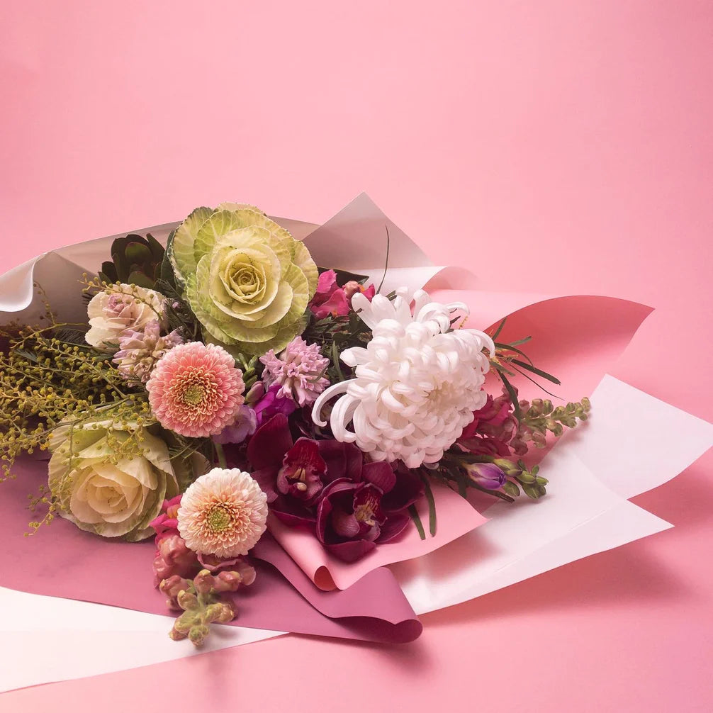 A vibrant bespoke floral bouquet of mixed flowers, including roses and chrysanthemums, artfully wrapped in paper against a pink background, from Poppy in April's Doreen Pretty & Pastel collection.