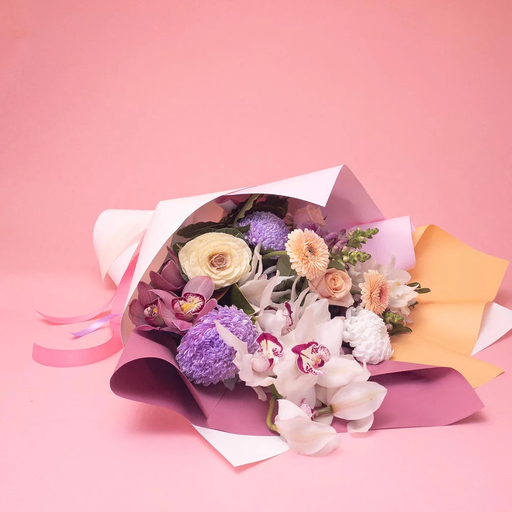 A delicately arranged bouquet of Doreen Pretty & Pastel flowers with purple accents, wrapped in white and yellow paper against a soft pink background, selected from Poppy in April's bespoke floral collections.