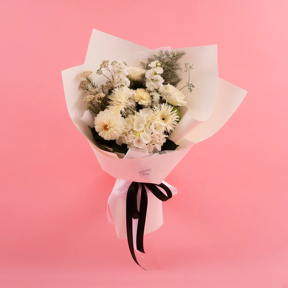 A bouquet of Doreen White & Green flowers from Poppy in April's bespoke floral collection, wrapped in white paper tied with a black ribbon, set against a pink background.
